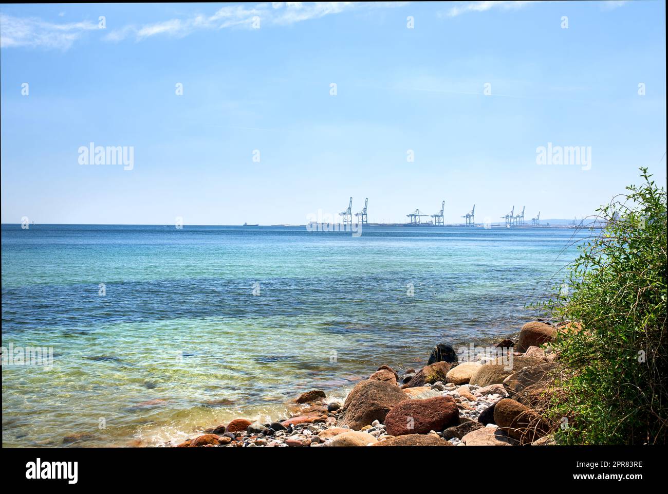 Copy space at sea with a blue sky background and harbour cranes at a port in the horizon. Calm ocean waves at a shallow and rocky shore. Scenic coastal landscape for a relaxing summer holiday Stock Photo
