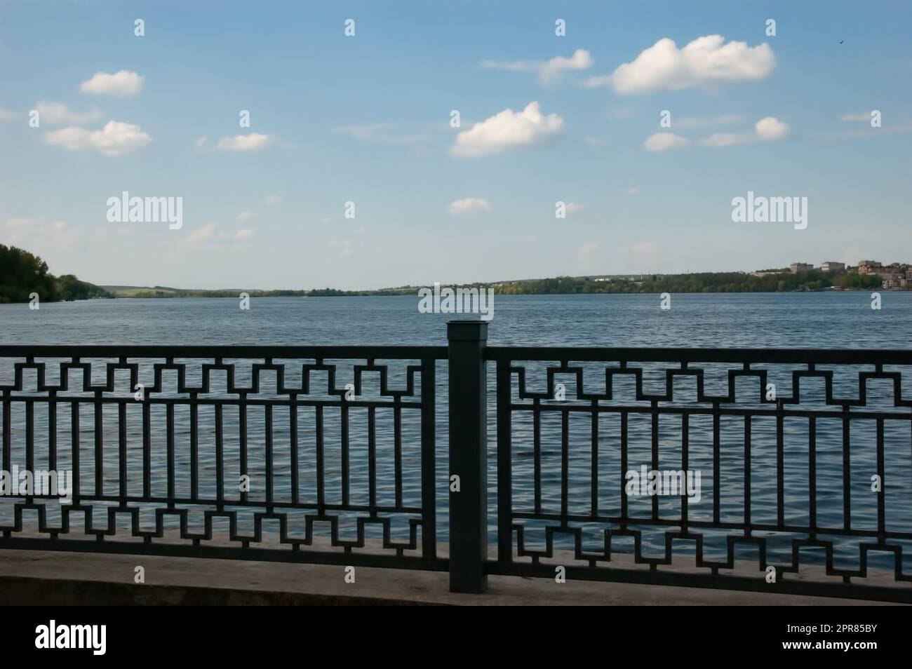 Tranquil lake behind a metal fence, blue sky with white clouds, houses in the distance Stock Photo