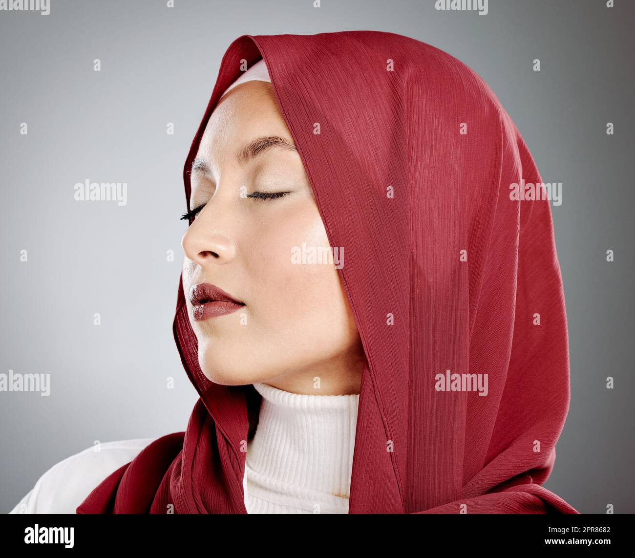 Stylish young muslim woman in a red hijab with her eyes closed on a grey studio background. Middle eastern woman wearing makeup and a headscarf while feeling calm and showing the beauty in modesty Stock Photo