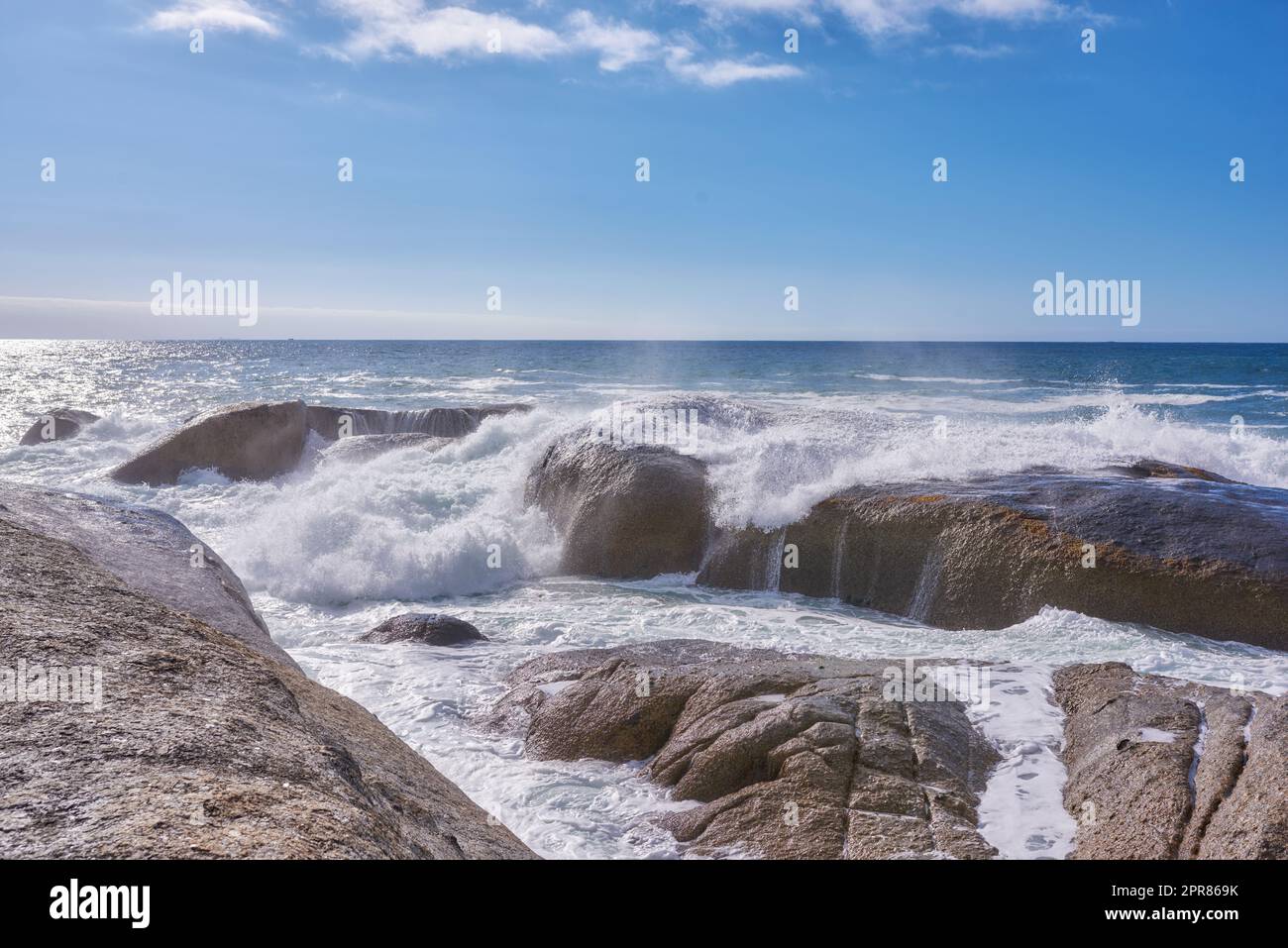 A rocky shore and a view of the ocean with waves, blue sky copy space, and a horizon in the background in Camps Bay, Cape Town, South Africa. Calm, serene, tranquil beach and nature scenery Stock Photo