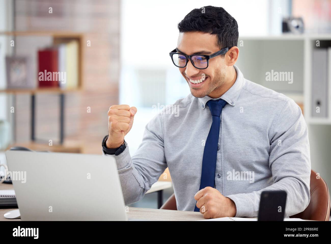 Young happy mixed race businessman cheering with his fist in joy while working on a laptop at work. Cheerful hispanic male businessperson celebrating success and victory in an office Stock Photo