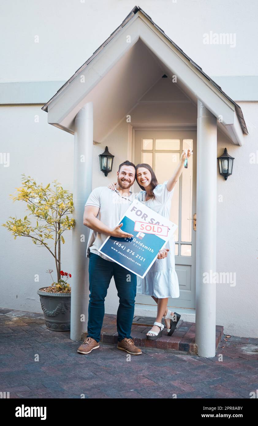 Young caucasian couple holding house keys while moving into new apartment. Happy man and woman holding for sale and sold sign outside home while relocating. Securing a loan for property or real estate Stock Photo