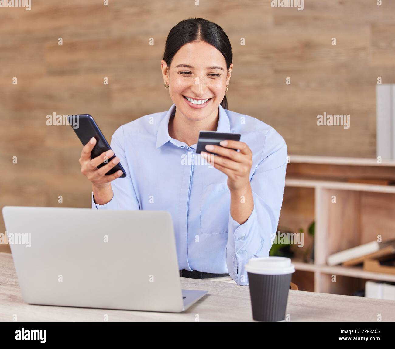 Beautiful young mixed race woman reading her credit card while using her phone and laptop to shop online while sitting in the office at work. Shopping has never been simpler or more convenient Stock Photo