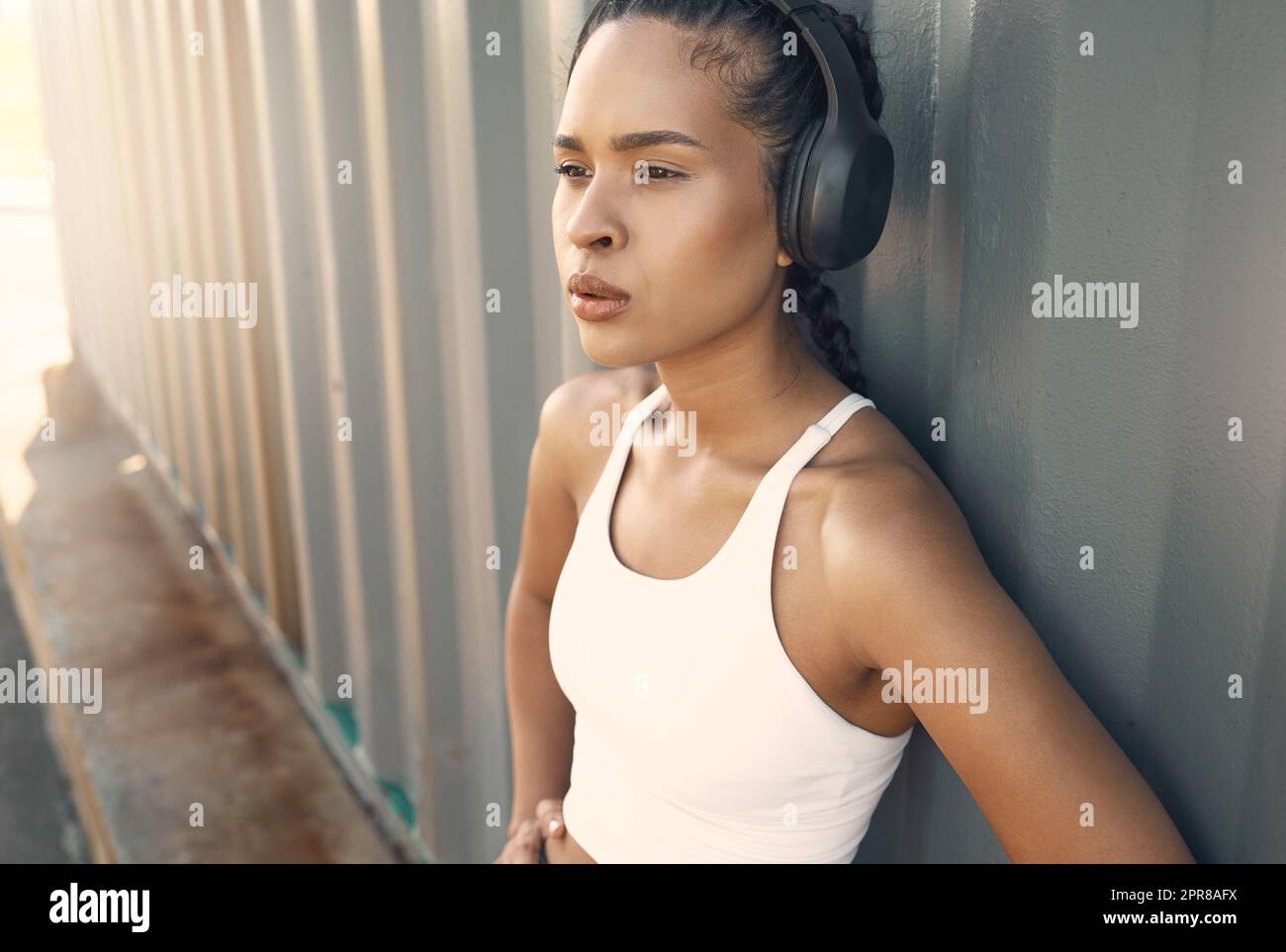 One fit young hispanic woman wearing headphones and taking a rest break to catch her breath after a run or jog in an urban setting outdoors. Female athlete looking tired after intense cardio exercise Stock Photo