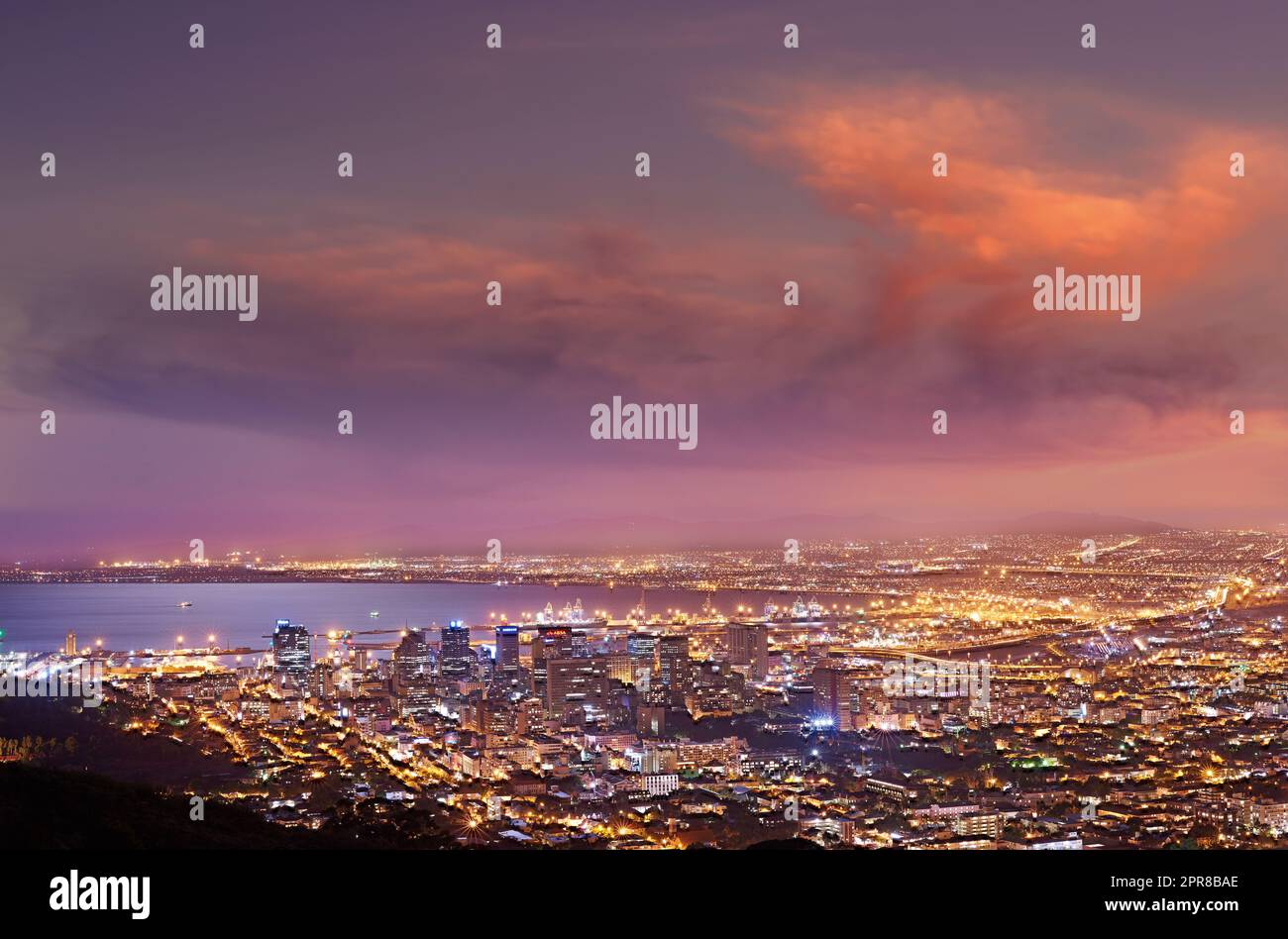City lights of Cape Town at sunset from above. Dreamy panoramic coastal urban landscape at night. Cityscape near a harbor on purple horizon with clouds. High angle view from Signal Hill, South Africa Stock Photo