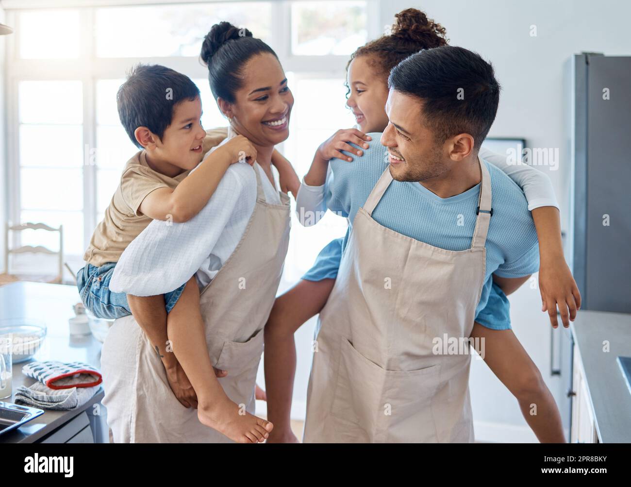 Energetic parents giving their children a piggyback ride while wearing aprons and baking in the kitchen. Hispanic mom and dad playing with kids at home Stock Photo