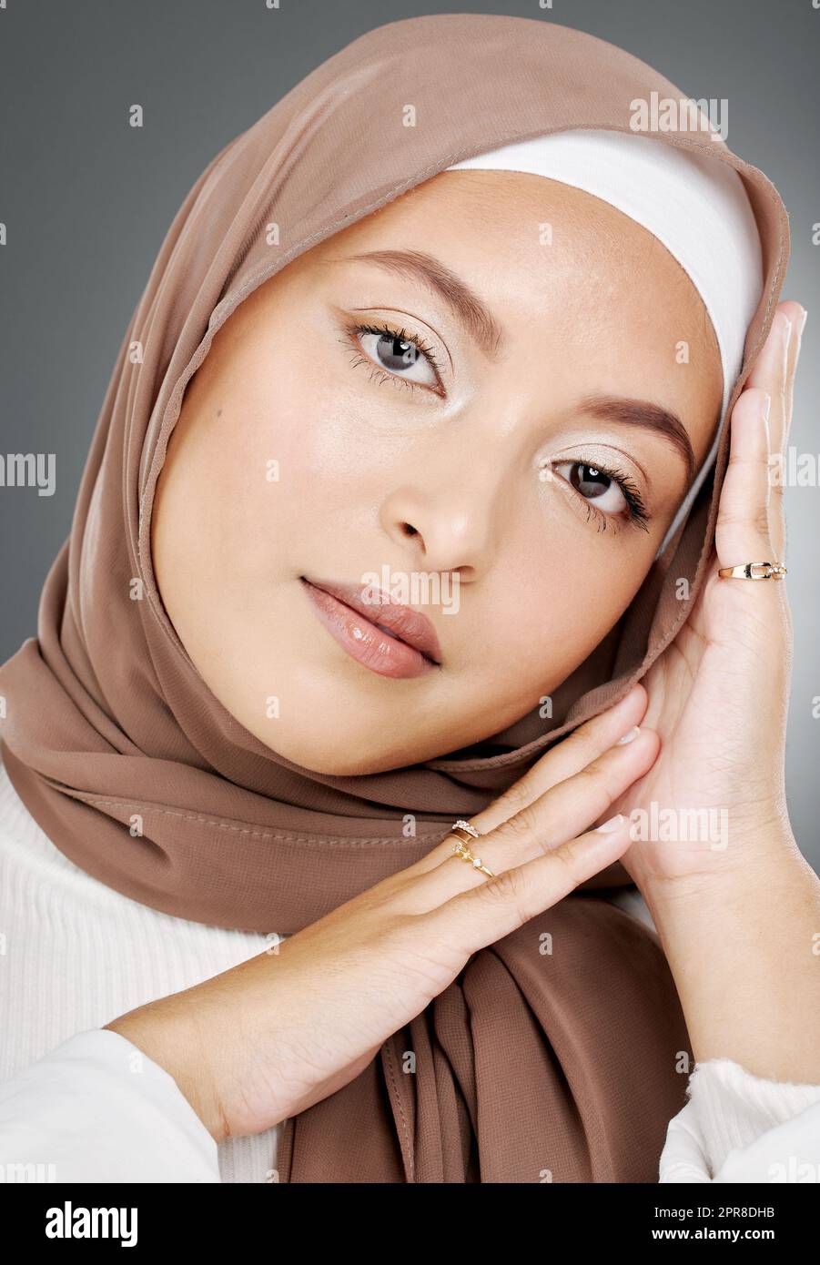 Portrait of a glowing elegant muslim womans face isolated against grey studio background. Young woman wearing a hijab or headscarf, showing her eyelash extensions, jewellery and skin routine Stock Photo
