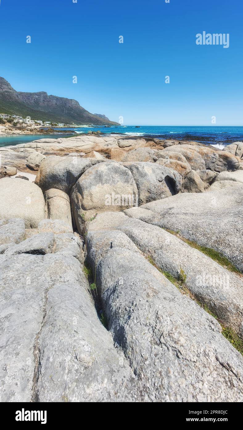 Rocky landscape leading to the sea under cloudy blue sky copy space at Camps Bay beach, Cape Town, South Africa. Boulders on coastal shore with the Twelve Apostles mountain in the background Stock Photo