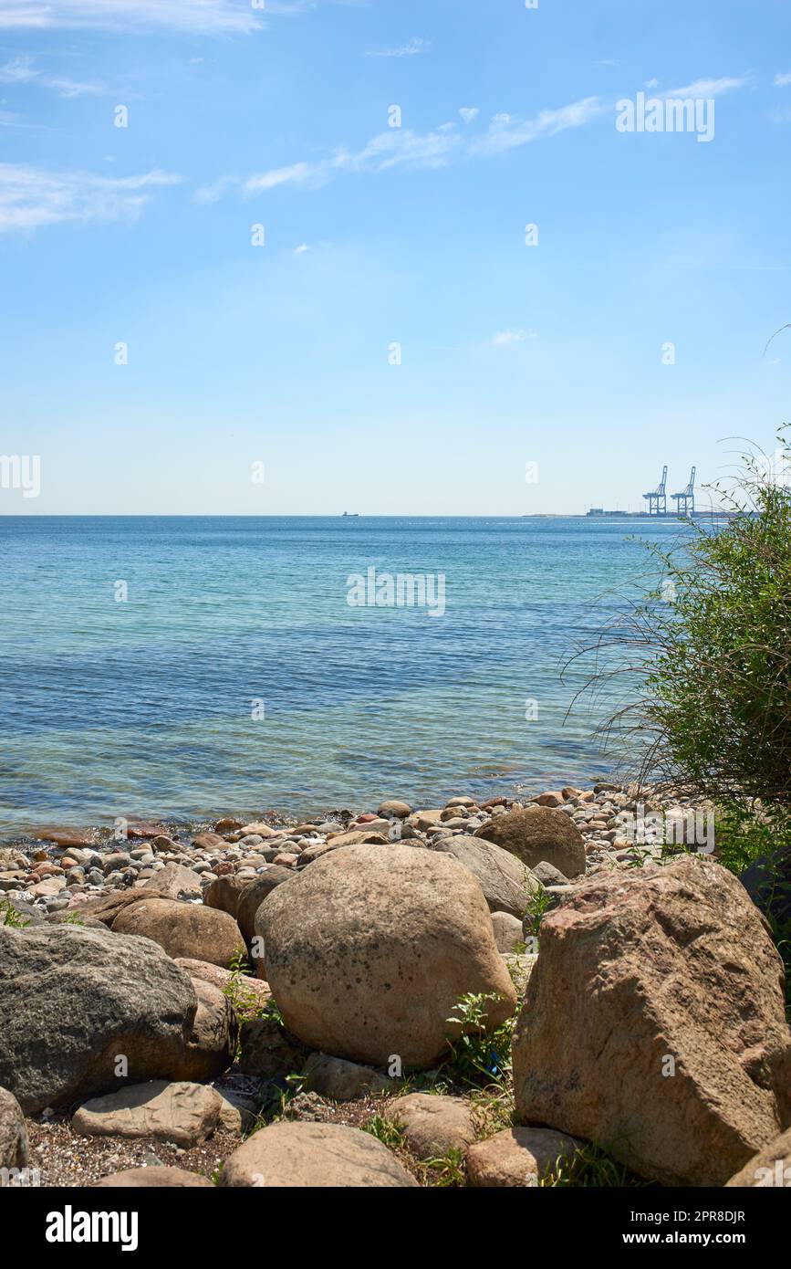 A calm quiet on the rocky beach coast of the Mediterranean ocean with the horizon and the intense illuminated blue sky with white clouds. A crystalline seascape with warm sunny weather during summer Stock Photo