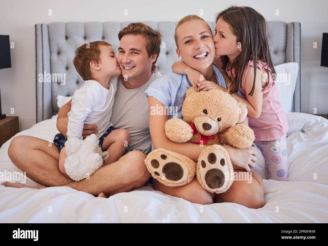 Loving children sharing tender moment with parents while gently kissing mom and dad on the cheek. Happy caucasian parents spending time with their little daughter and son at home in the morning Stock Photo