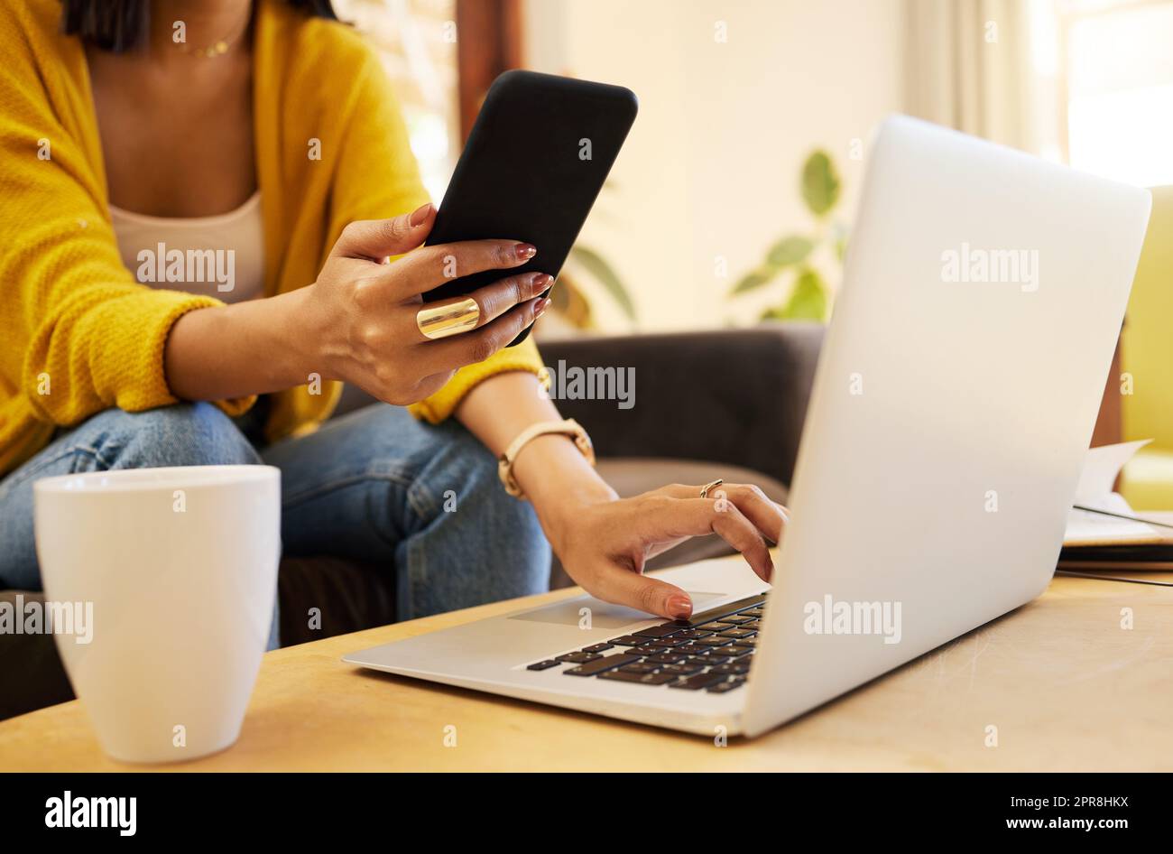 Closeup of woman working remote while typing on her laptop and holding her smartphone sitting on a sofa in a bright living room. One focused hispanic young female at home using modern technology Stock Photo