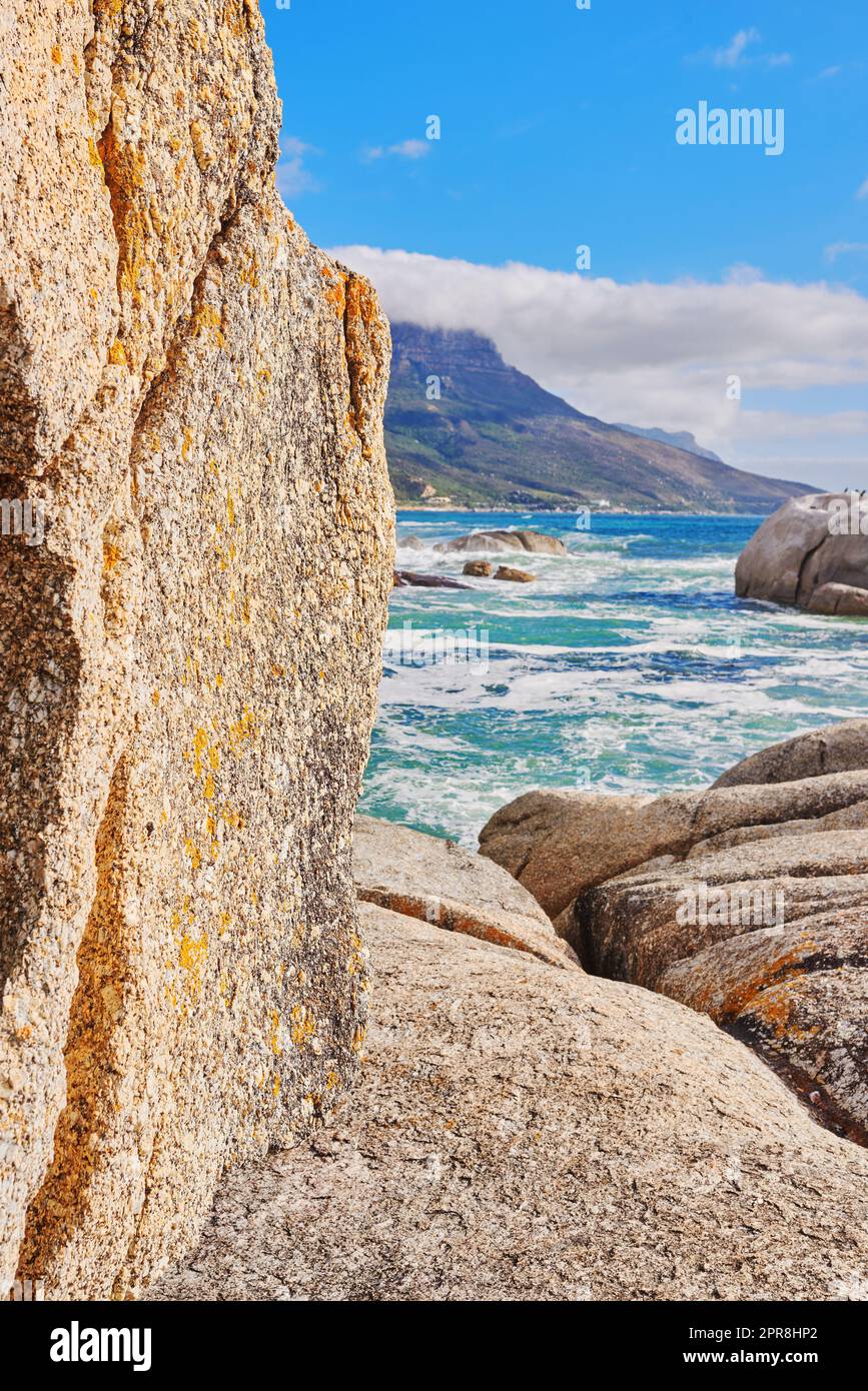 Stunning seaside location for a summer holiday in Cape Town. Boulders at a beach with ocean waves and water washing over rocks at the coast with mountain and blue sky with clouds in the background Stock Photo