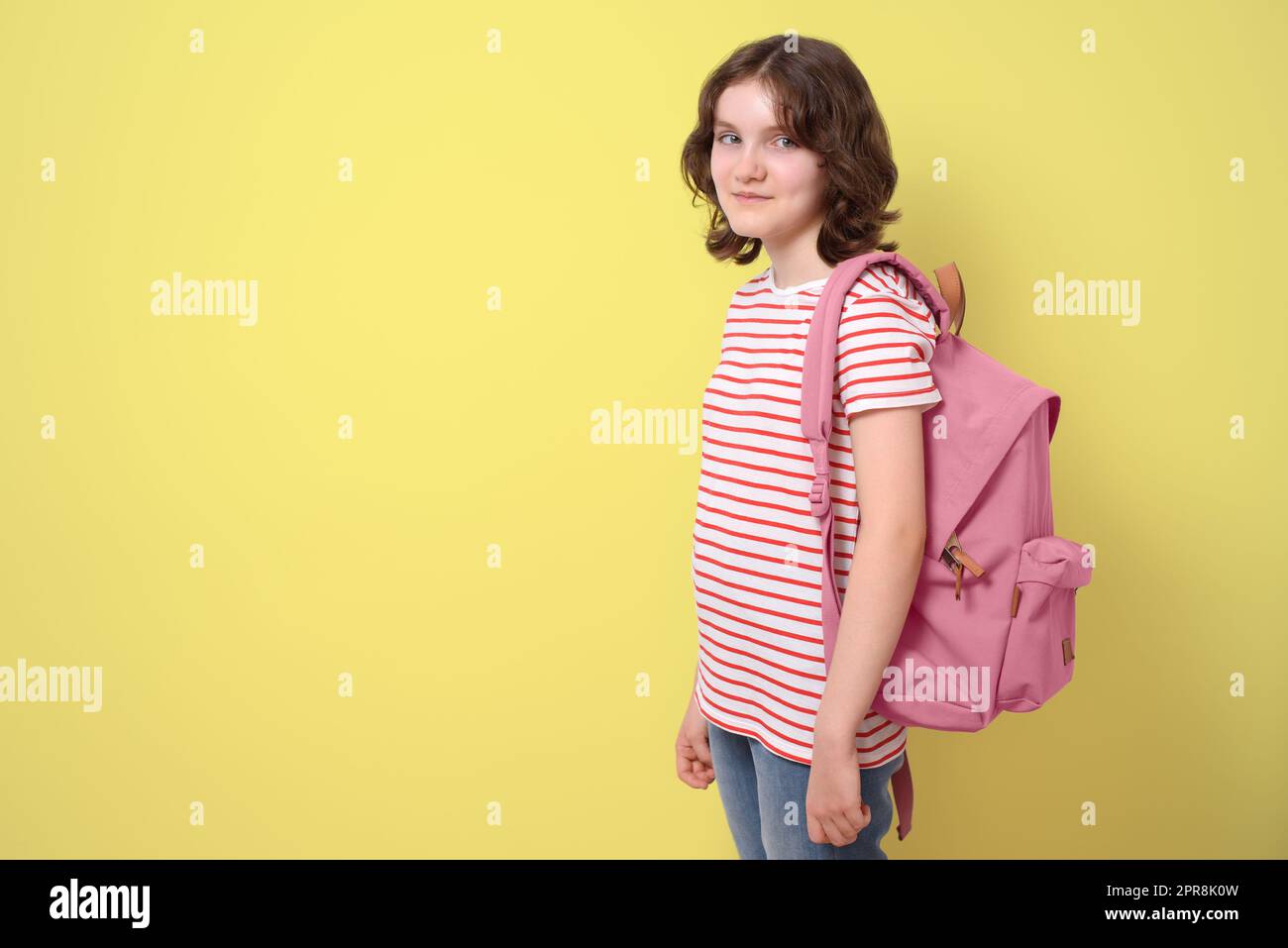 Schoolgirl standing, smiling and holding a backpack, looking at camera, on yellow background Stock Photo