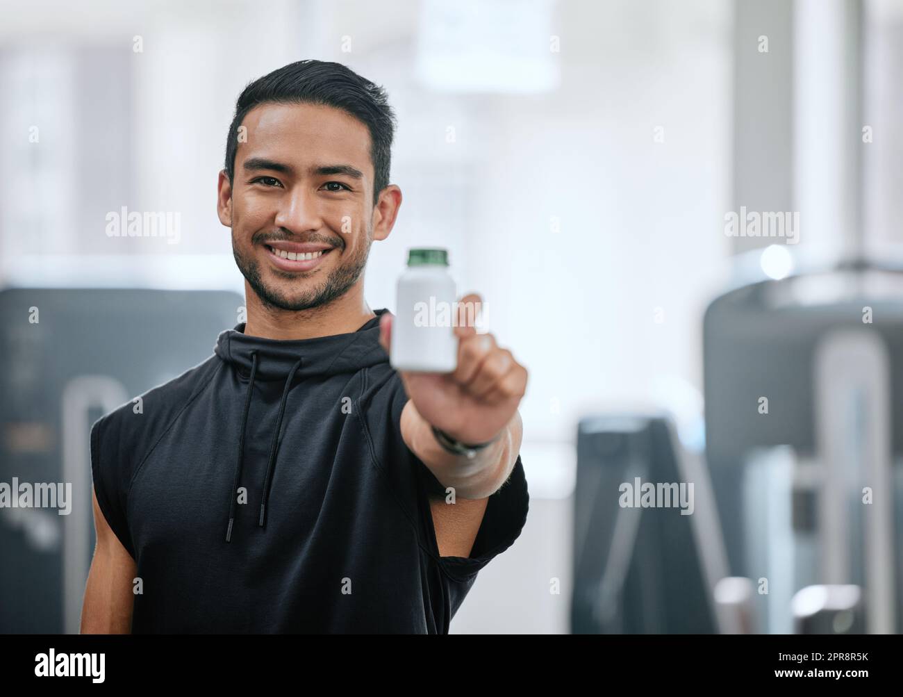 Portrait of smiling trainer alone in gym, holding and showing bottle of steroid pills. Asian coach with hormone enhancing drugs for workout in exercise health club. Bodybuilder man in fitness centre Stock Photo