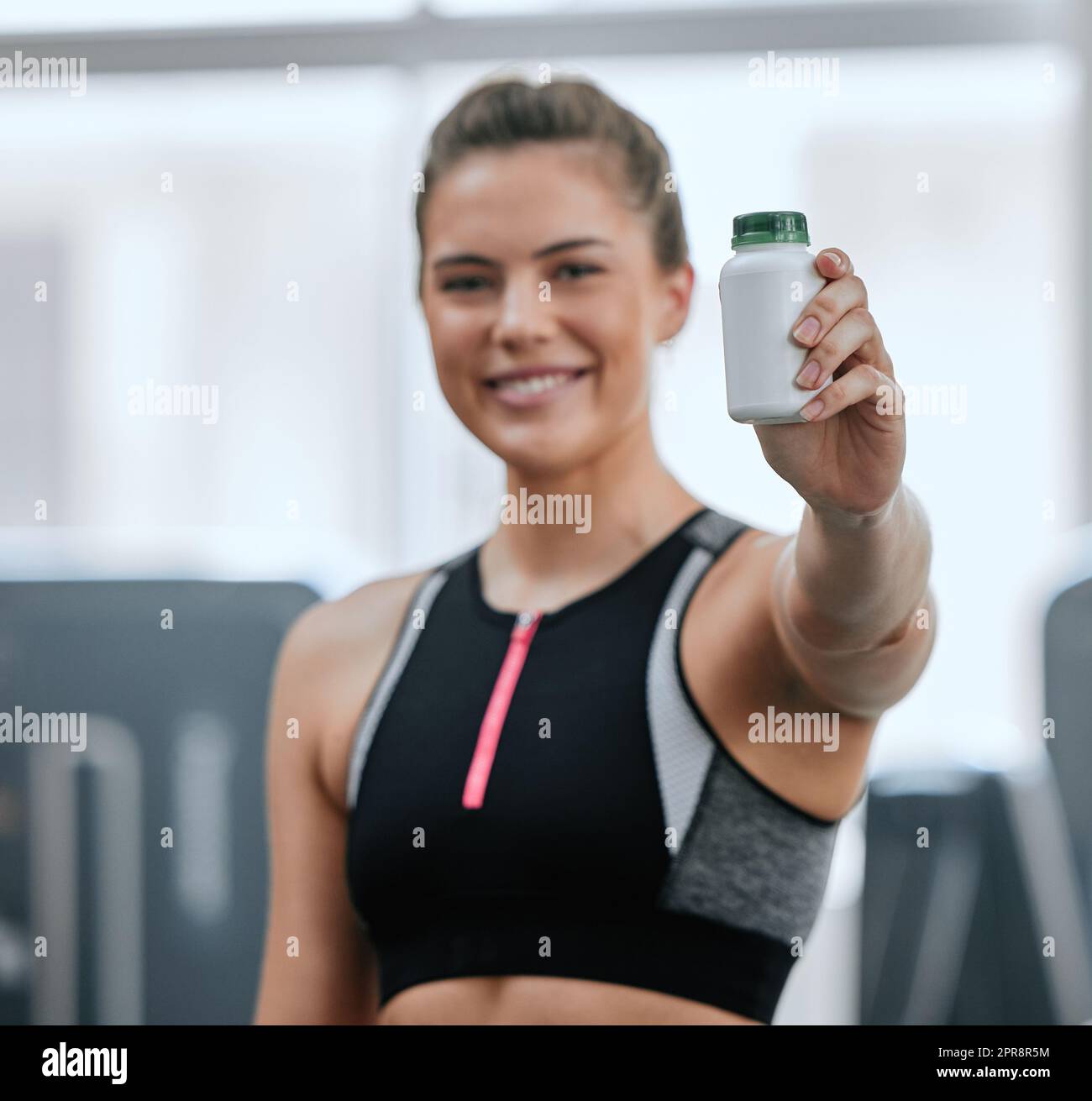 Smiling trainer alone in gym while holding and showing bottle of steroid pills. Caucasian coach with hormone enhancing drugs for workout in exercise health club. Bodybuilder woman in fitness centre Stock Photo