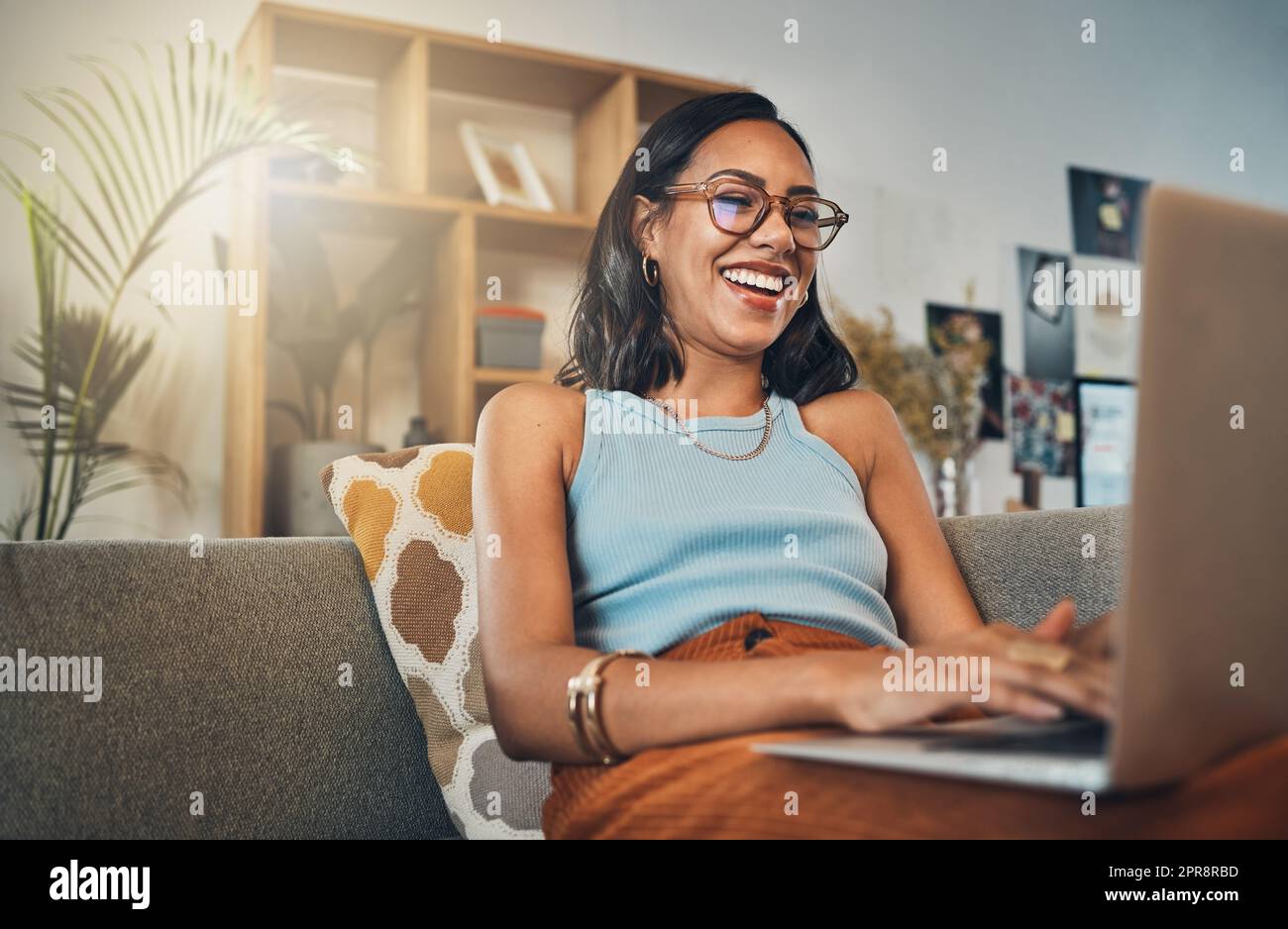 Smiling mixed race woman browsing internet on a laptop at home. One happy hispanic entrepreneur sitting alone on her sofa and blogging over a weekend. Relaxing in a living room and typing a blog post Stock Photo