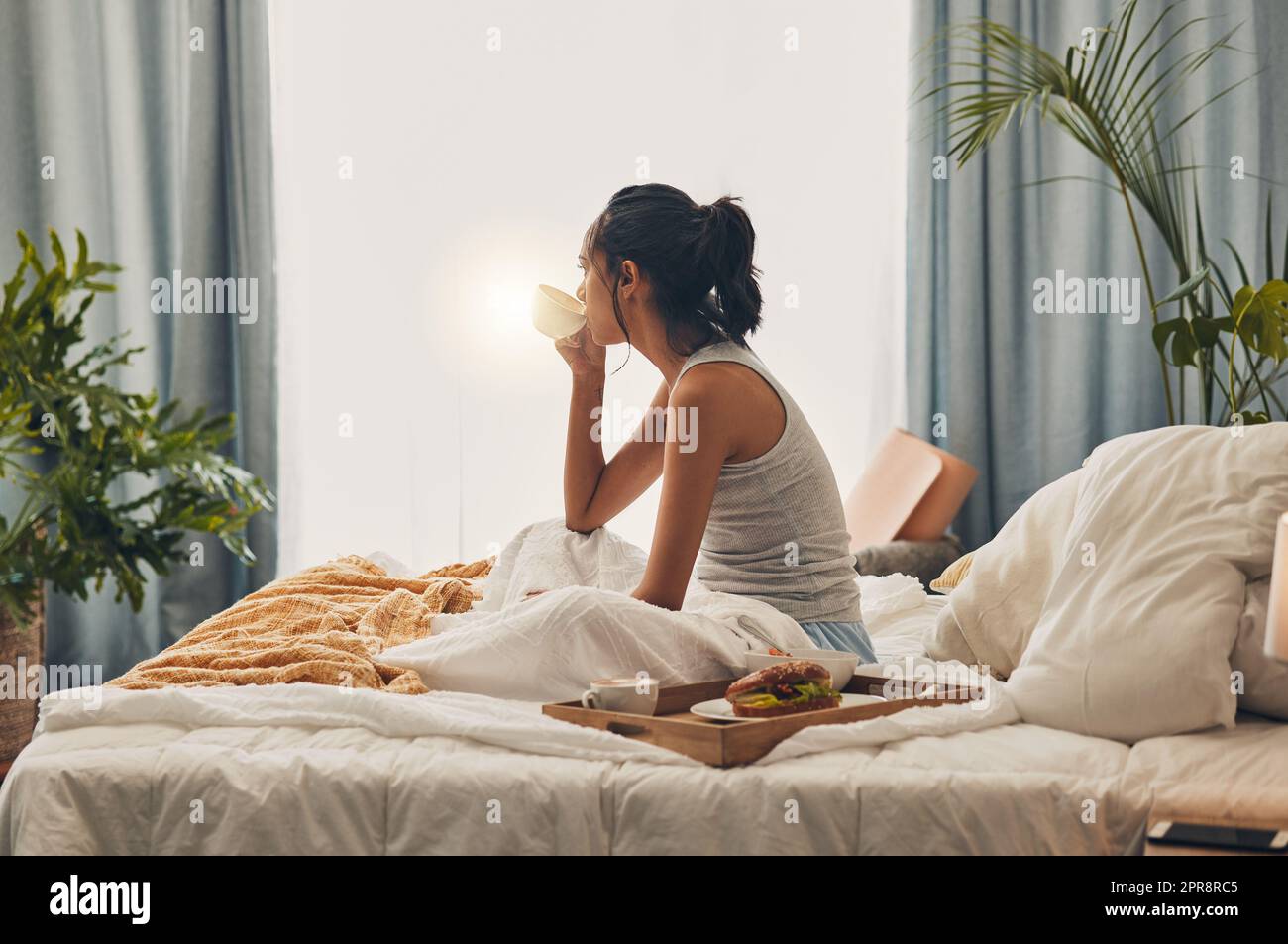 A beautiful young Hispanic woman enjoying a warm cup of coffee for breakfast. One mixed race female drinking tea while sitting in bed and daydreaming Stock Photo