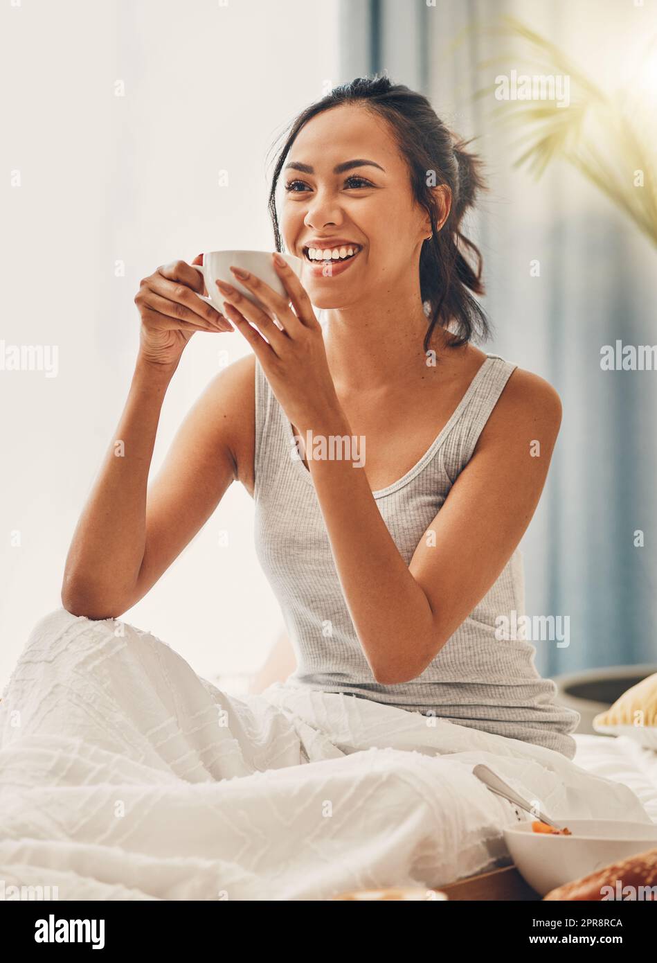 A beautiful young Hispanic woman enjoying a warm cup of coffee for breakfast. One mixed race female drinking tea while sitting in bed and laughing Stock Photo