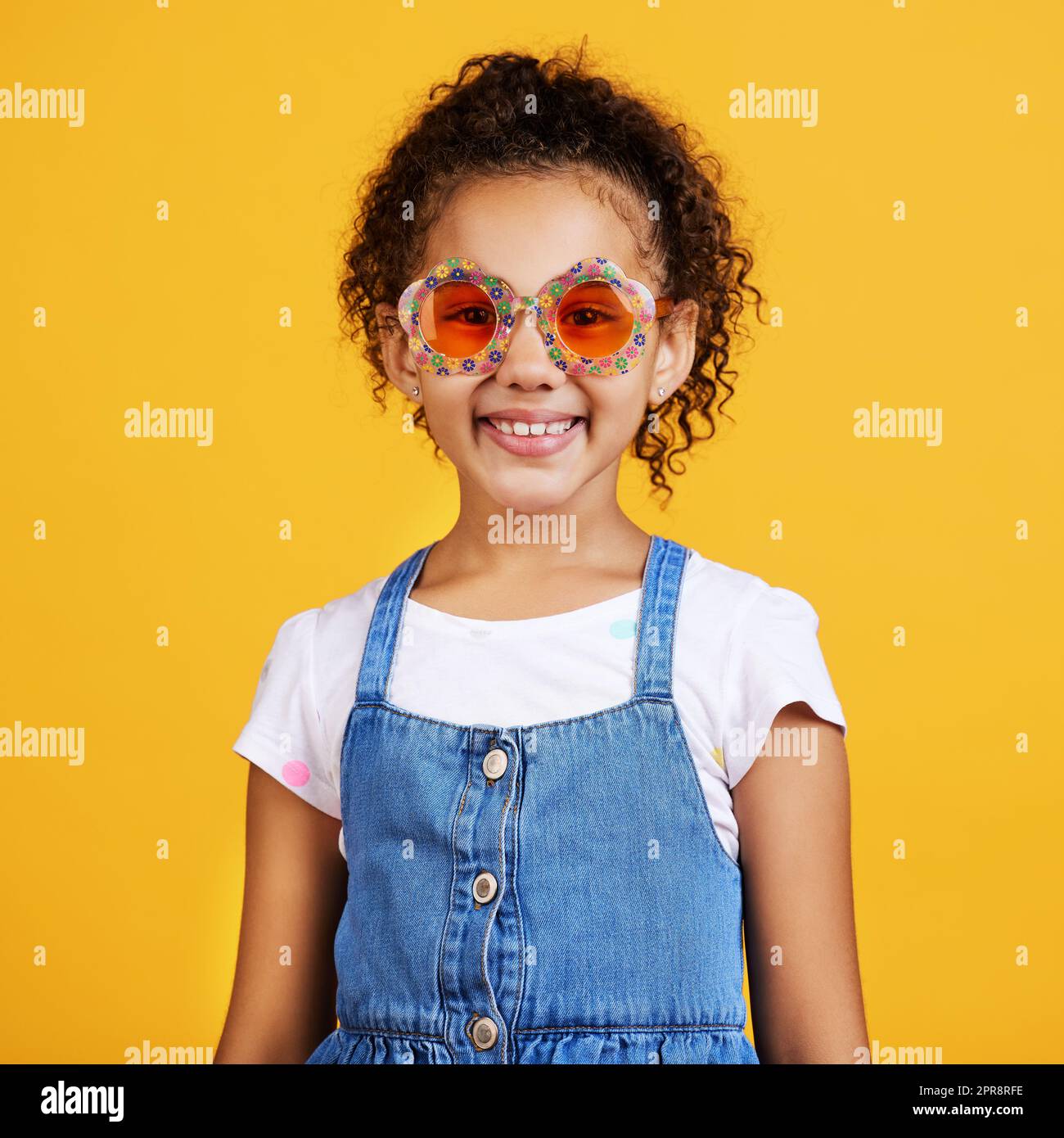 Studio portrait mixed race girl wearing funky sunglasses Isolated against a yellow background. Cute hispanic child posing inside. Happy and carefree kid with an imagination for being a fashion model Stock Photo