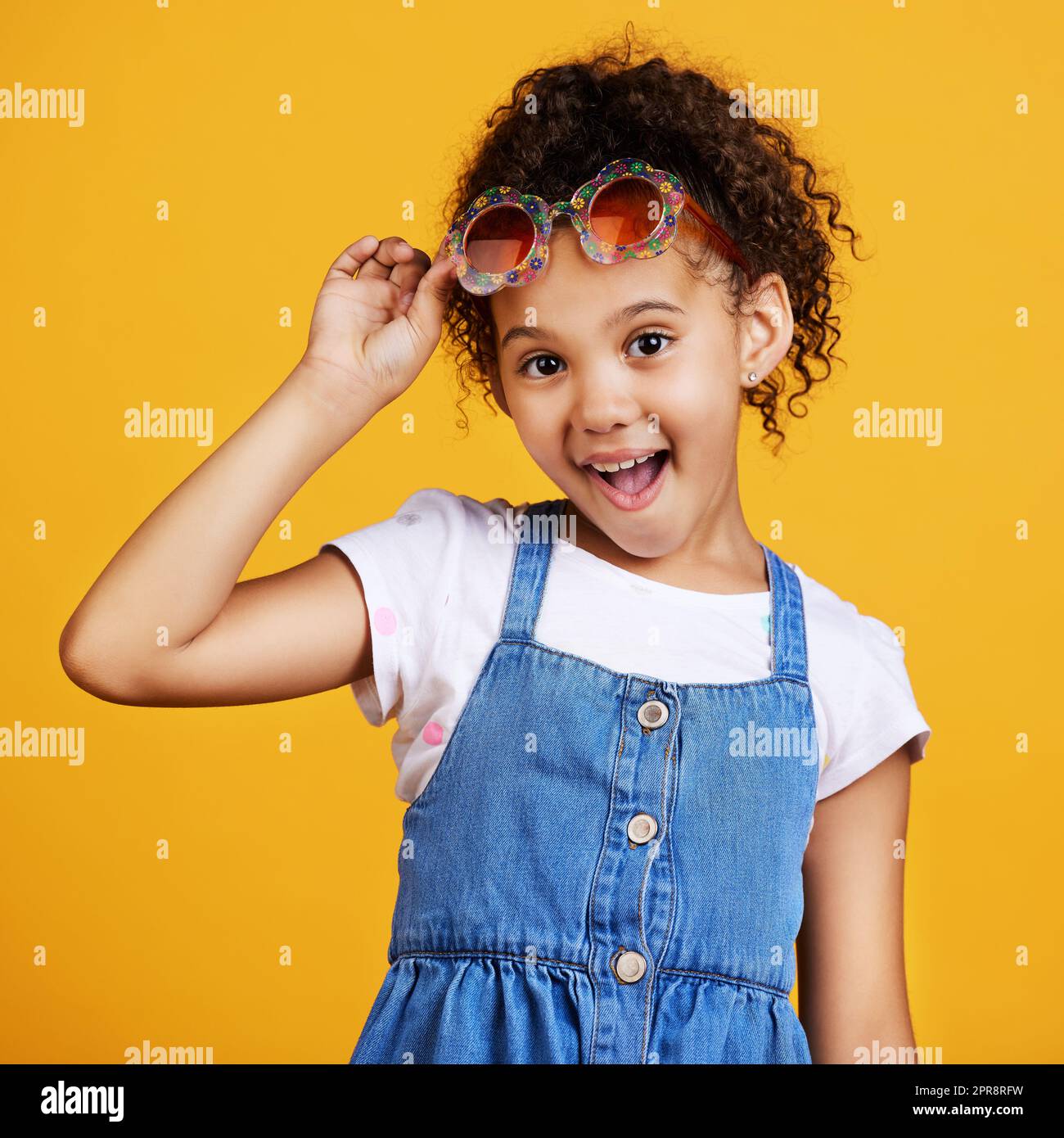 Studio portrait mixed race girl wearing funky sunglasses Isolated against a yellow background. Cute hispanic child posing inside. Happy and carefree kid with an imagination for being a fashion model Stock Photo