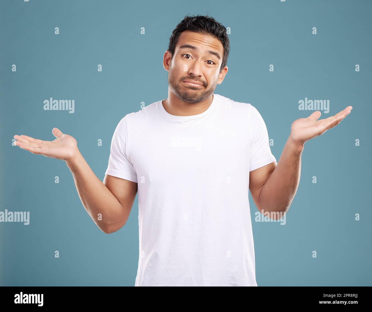 Handsome young mixed race man shrugging his shoulders while standing in studio isolated against a blue background. Confused hispanic male looking lost or clueless and making a, So what gesture Stock Photo