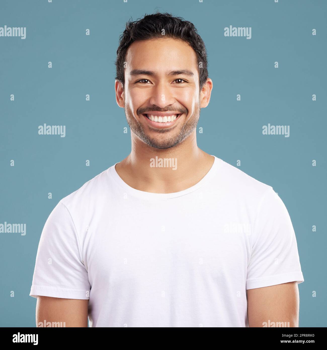 Handsome young mixed race man smiling and happy while standing in studio isolated against a blue background. Hispanic male in casual wear expressing happiness with a smile and looking at the camera Stock Photo