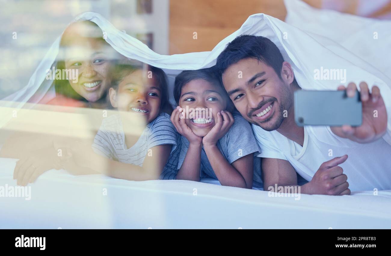 Happy parents with little children lying on bed at home under blanket and taking selfies on cellphone. Smiling mixed race girls bonding and enjoying free time with their mother and father Stock Photo