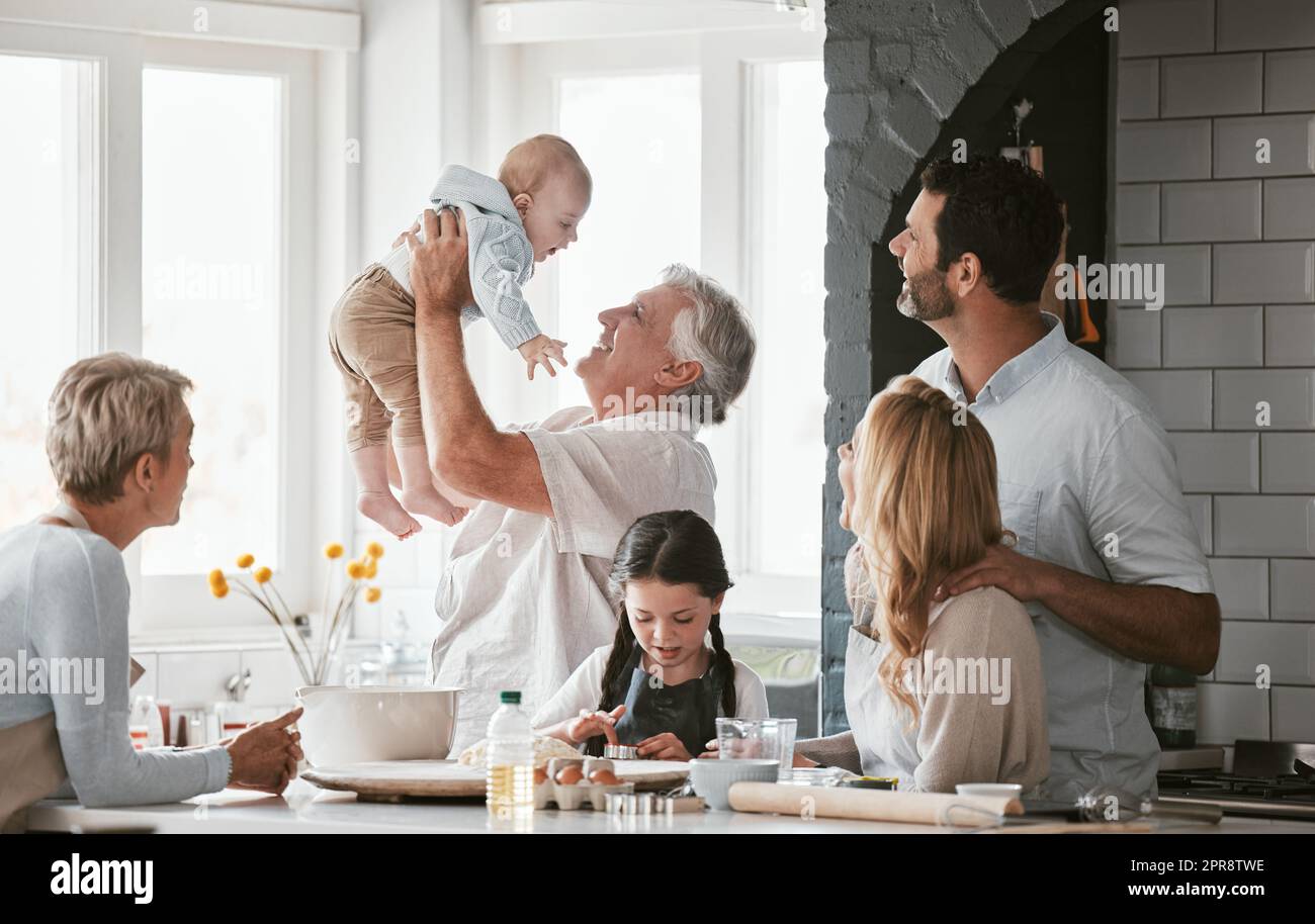 . a multi generation caucasian family bonding while spending the day together at the kitchen table at home. Grandparents being playful while holding the newborn baby. Young couple with two kids. Stock Photo