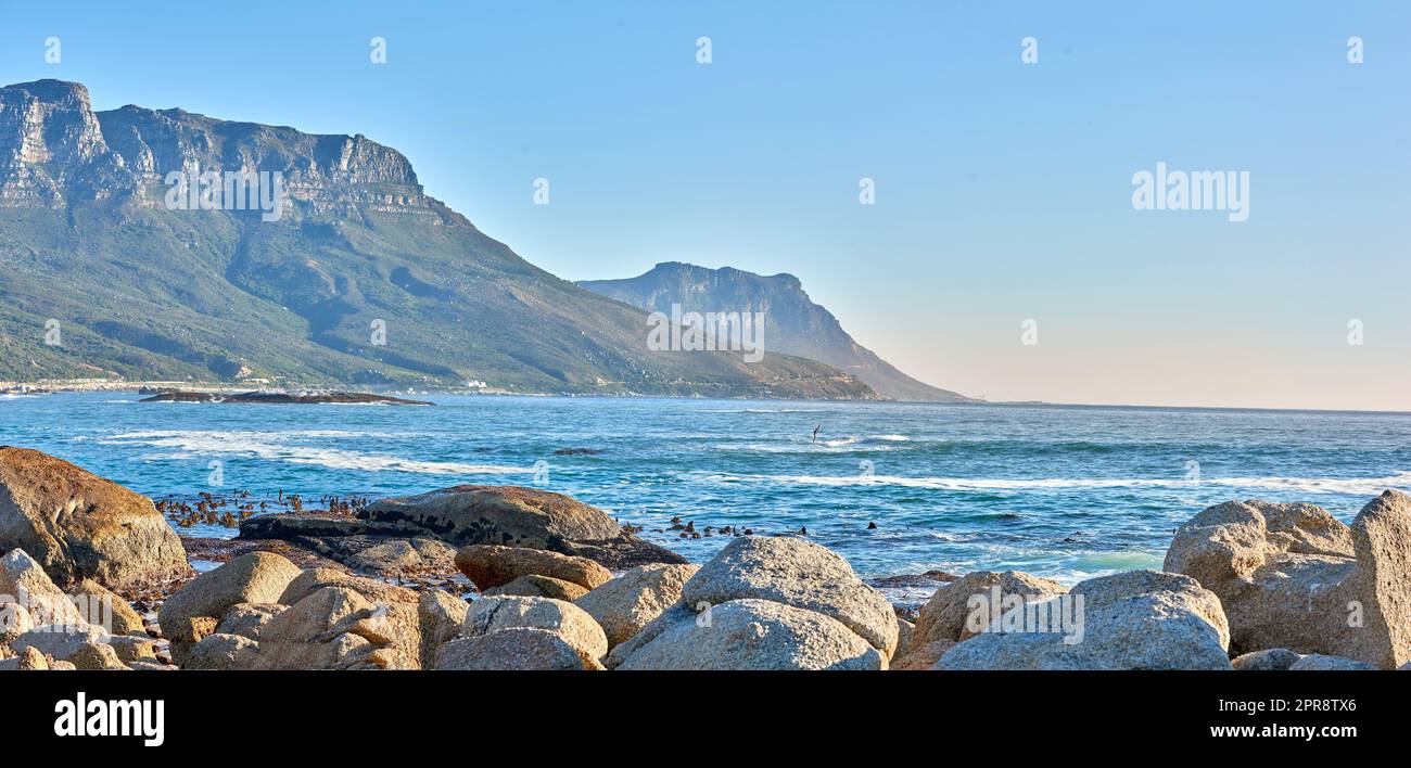 Quiet and calm ocean with rocky shore and boulders. Scenic seascape of the Twelve Apostles mountain under a blue sky in Cape Town. A beautiful holiday destination for travel and tourism Stock Photo