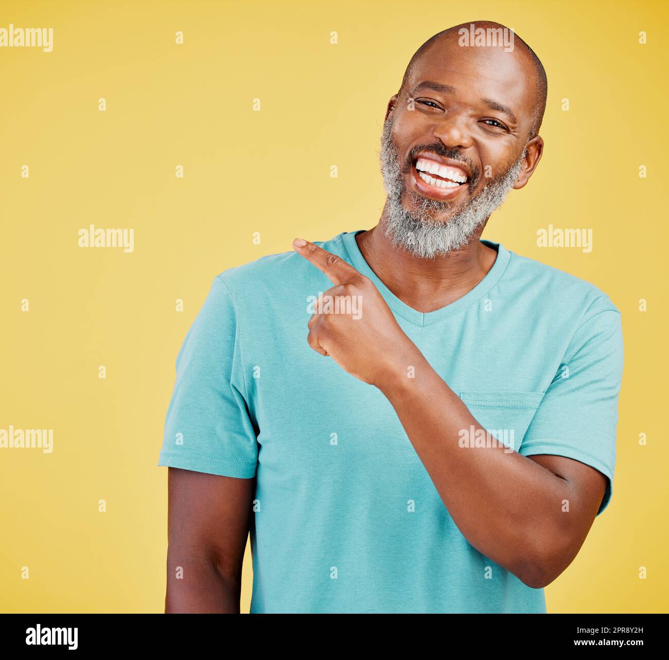 Mature african man laughing and pointing in a direction against a yellow studio background. Black guy giggling and making a pointing gesture reacting with laughter while looking cheerful and happy. Lighten the mood with an amusing and funny joke Stock Photo