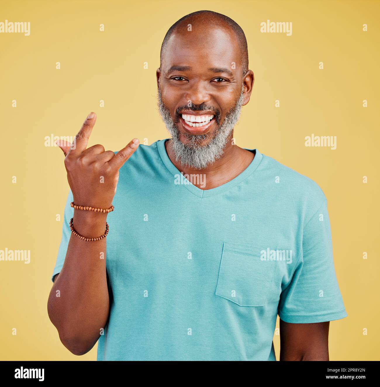 Happy mature african man expressing a rock and roll symbol gesture with his hands against a yellow studio background. Carefree, cool and confident black man making horn gesture to show good vibes Stock Photo