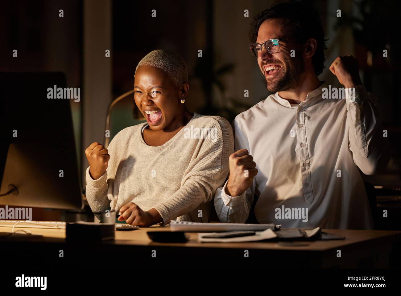 We scored big time. two businesspeople cheering while working on a computer in an office at night. Stock Photo