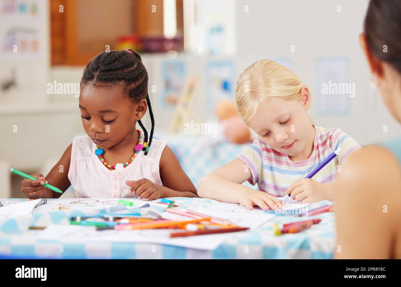 African american girl sitting at a table and colouring at pre-school or kindergarten with her caucasian classmate . Young female children using colourful pencils to draw pictures in a class at school Stock Photo