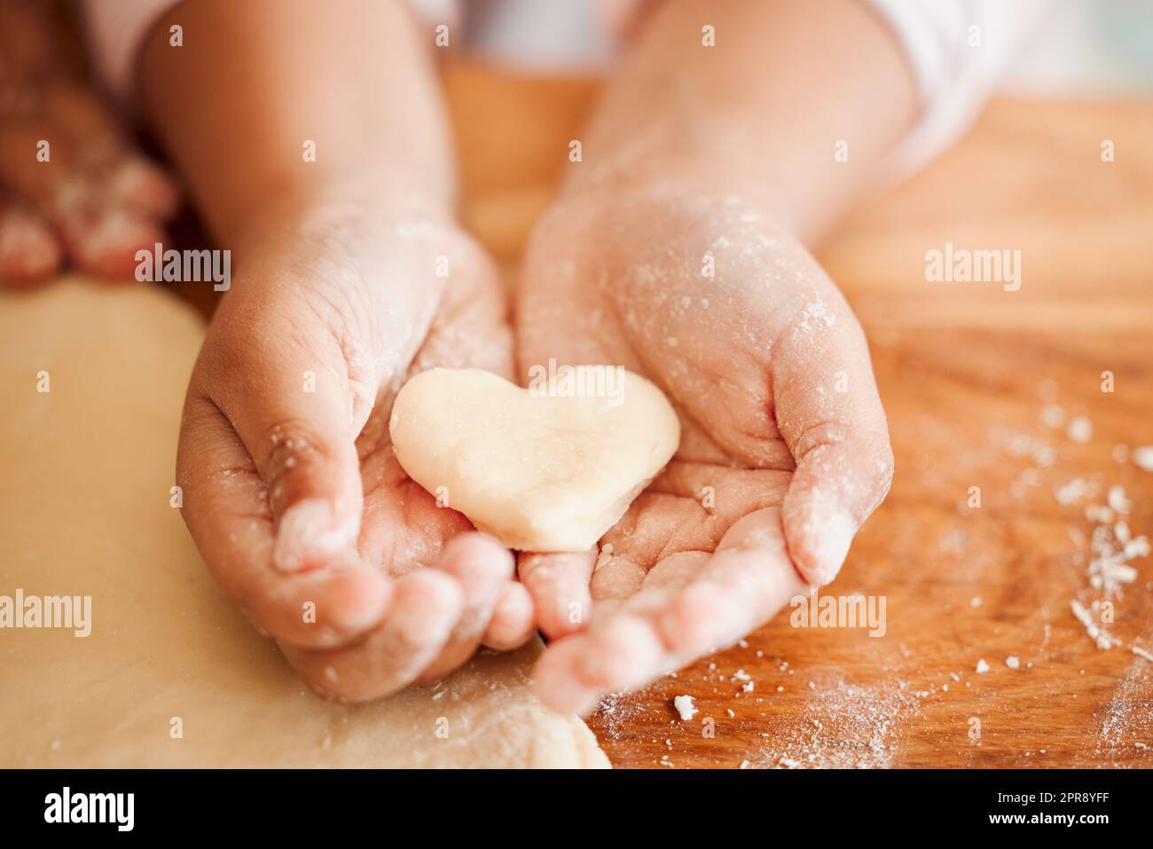 Closeup of an unknown chid holding a heart shaped cookie while baking in the kitchen with her mother. An unrecognizable mother teaching her daughter domestic skills at home Stock Photo