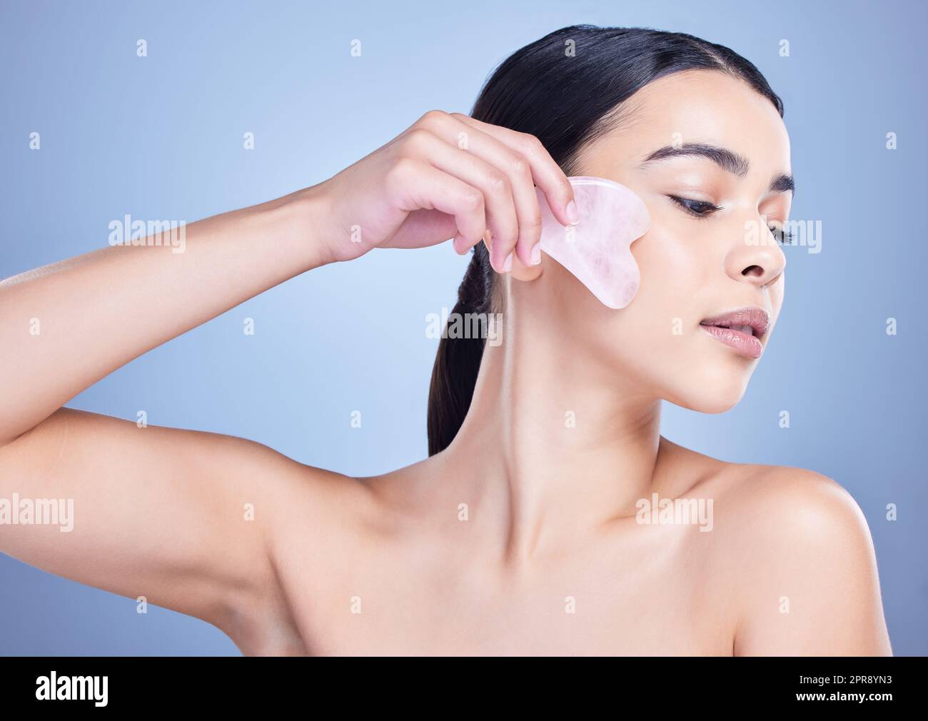 A beautiful mixed race woman using a rose quartz gua sha to reduce wrinkles and promote cell renewal. Young hispanic woman using anti ageing tool against blue copyspace background Stock Photo