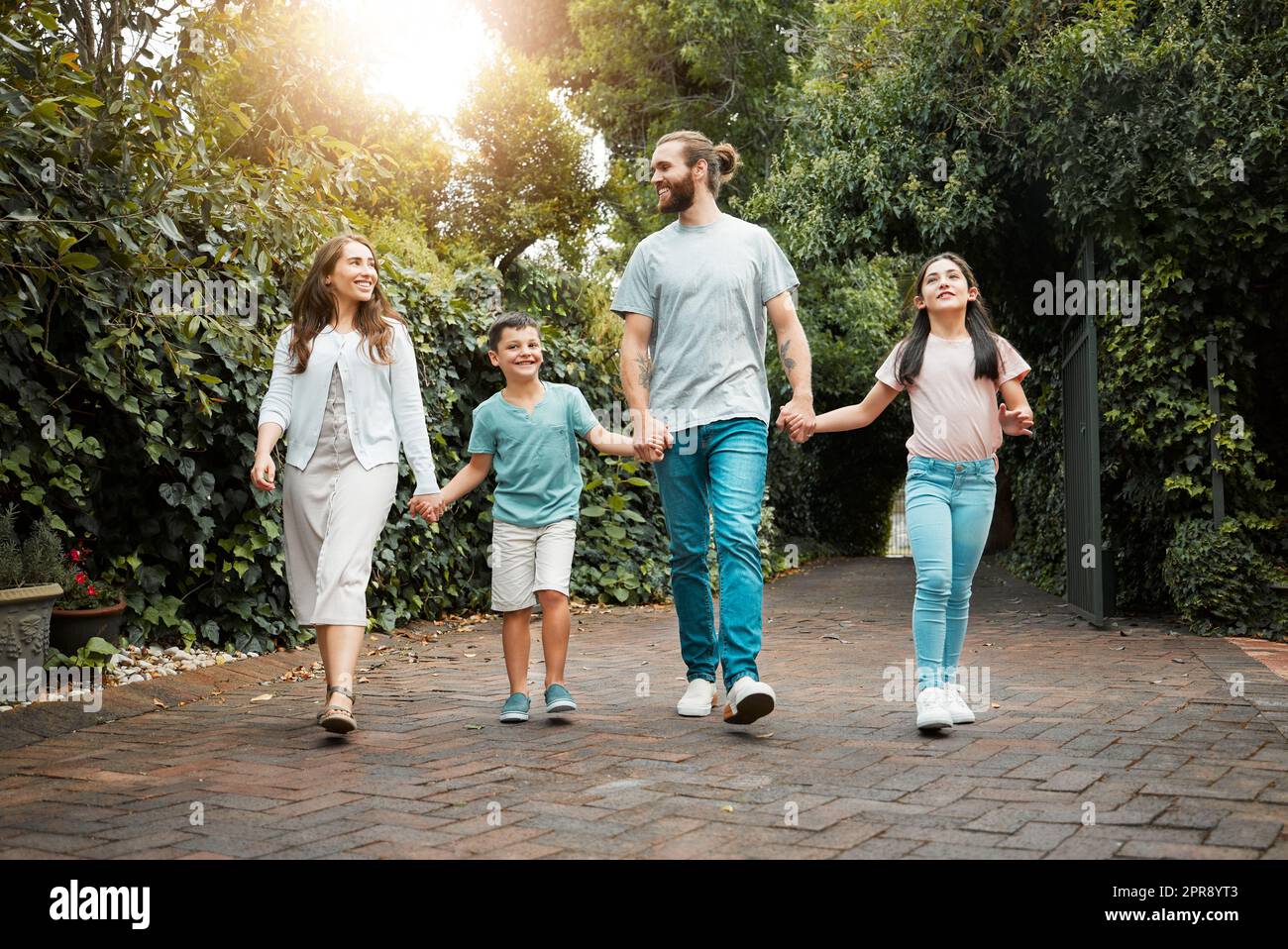 Happy young caucasian family of four holding hands and going for a walk in the neighbourhood. Carefree parents with their two children leaving their residence and going for a sunny stroll Stock Photo