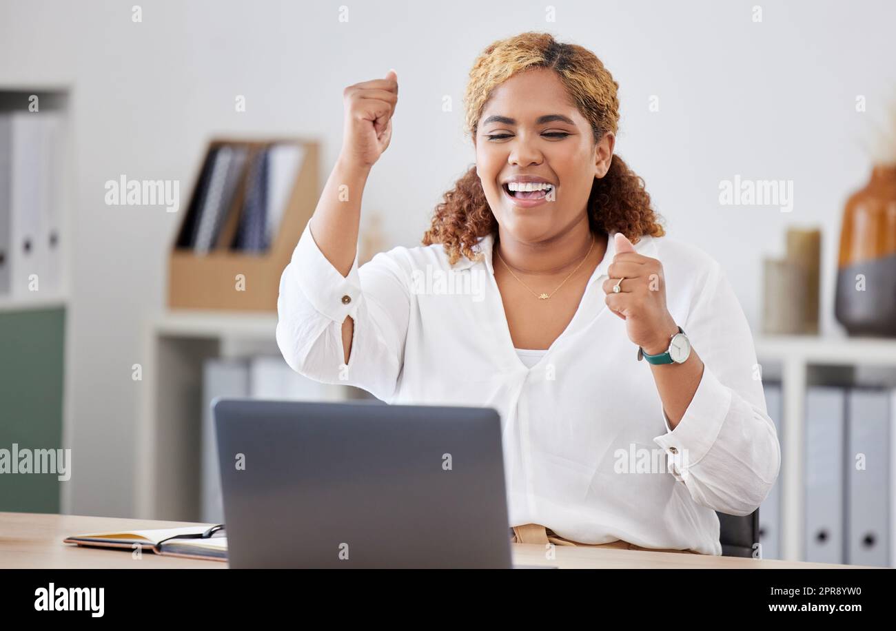Mixed race businesswoman cheering in joy while using a laptop alone at work. One hispanic businesswoman looking excited while working on a computer at a desk in an office Stock Photo