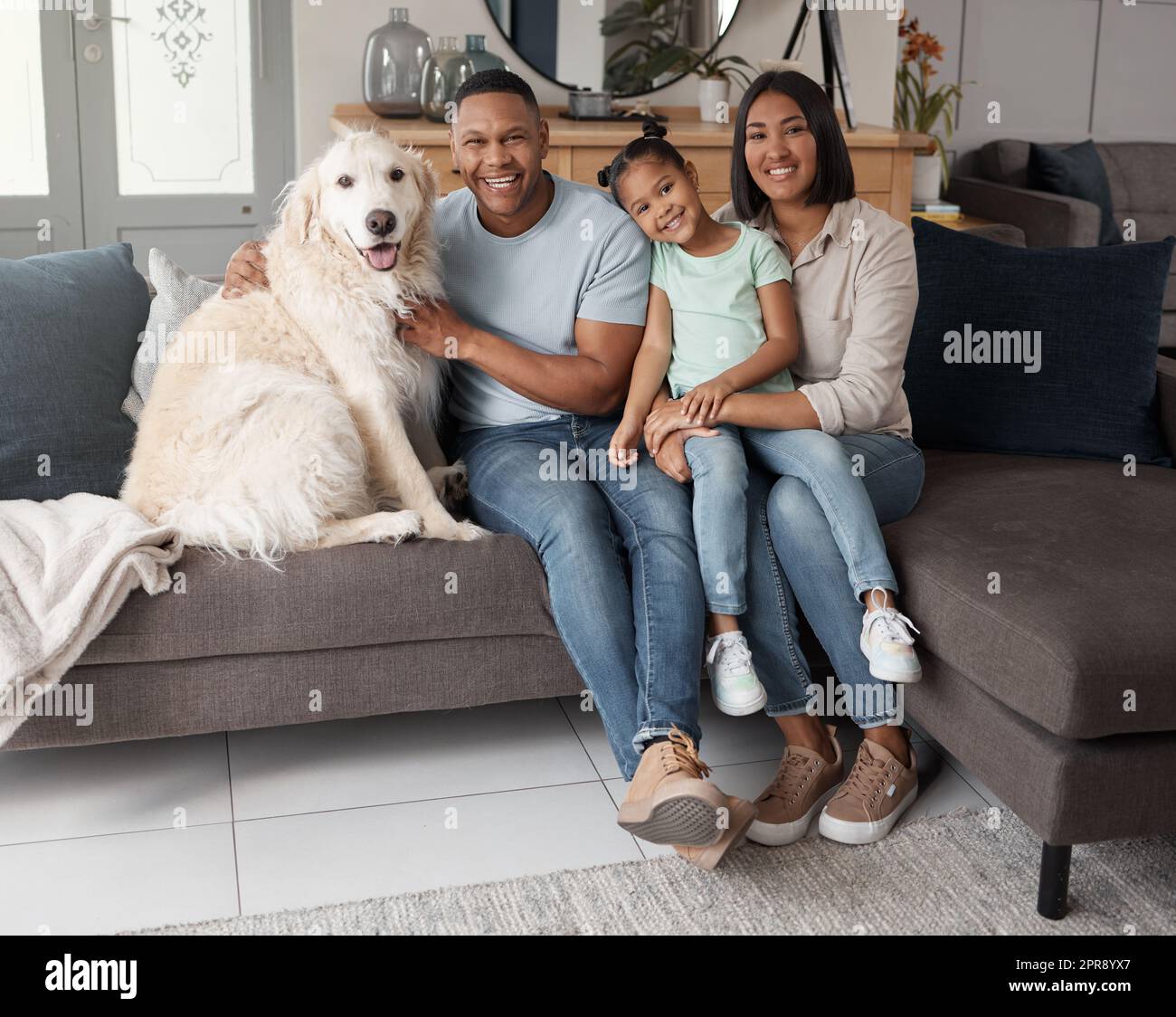 Portrait of a happy mixed race family of three relaxing on the sofa with their dog. Loving black family being affectionate with a foster animal. Young couple bonding with their daughter and rescued puppy at home Stock Photo