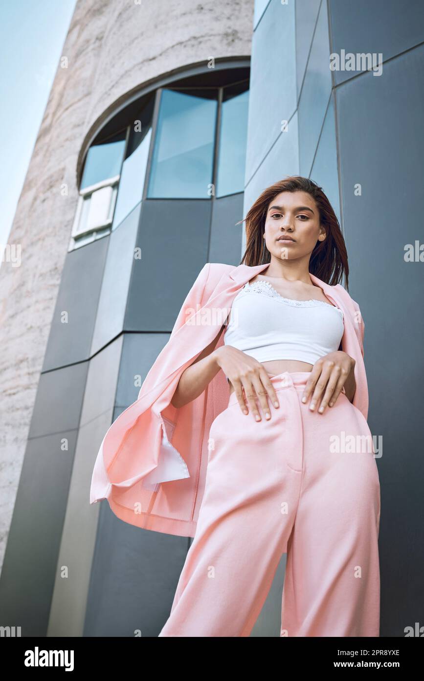 Portrait of a young trendy and confident mixed race woman looking stylish while posing and chilling time in the city. Fashionable hispanic woman wearing pink clothes and standing outside downtown Stock Photo