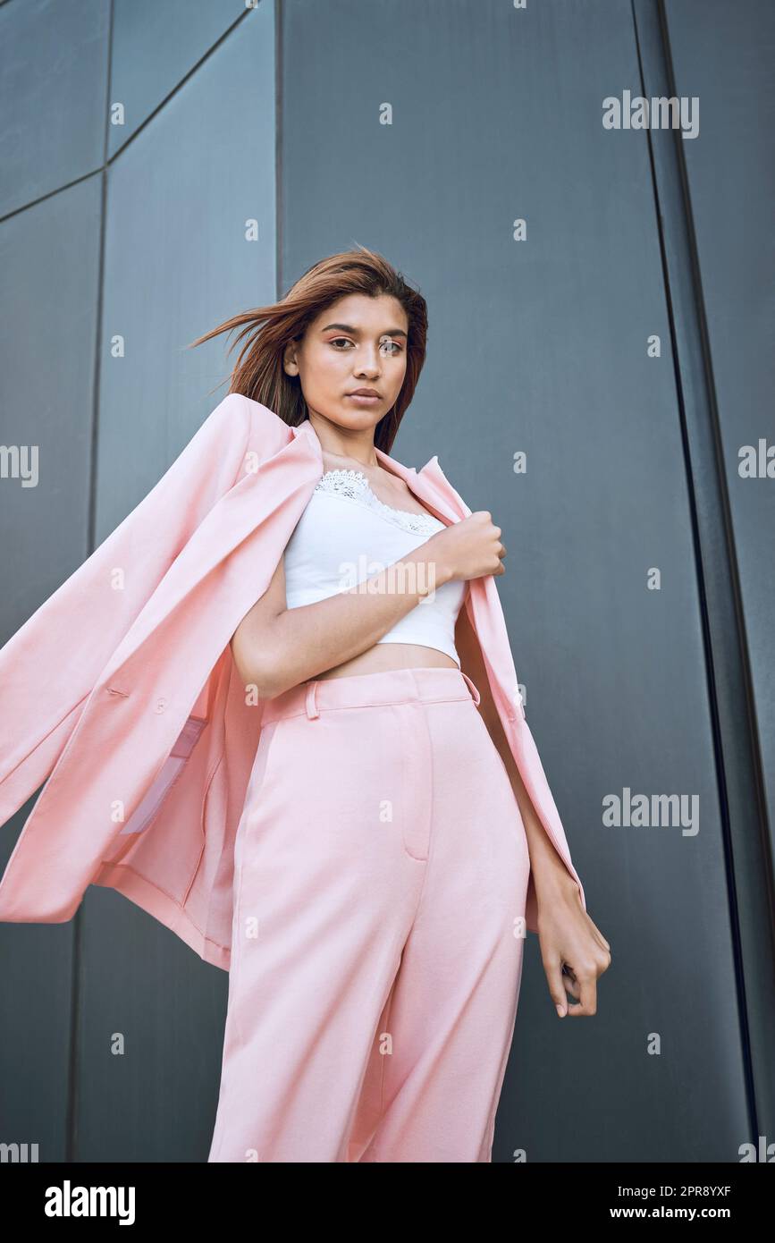 Below view portrait of young beautiful mixed race woman standing alone in a city and posing in fashionable clothes. Serious hispanic in trendy pink suit showing street style. Feeling bold, confident Stock Photo