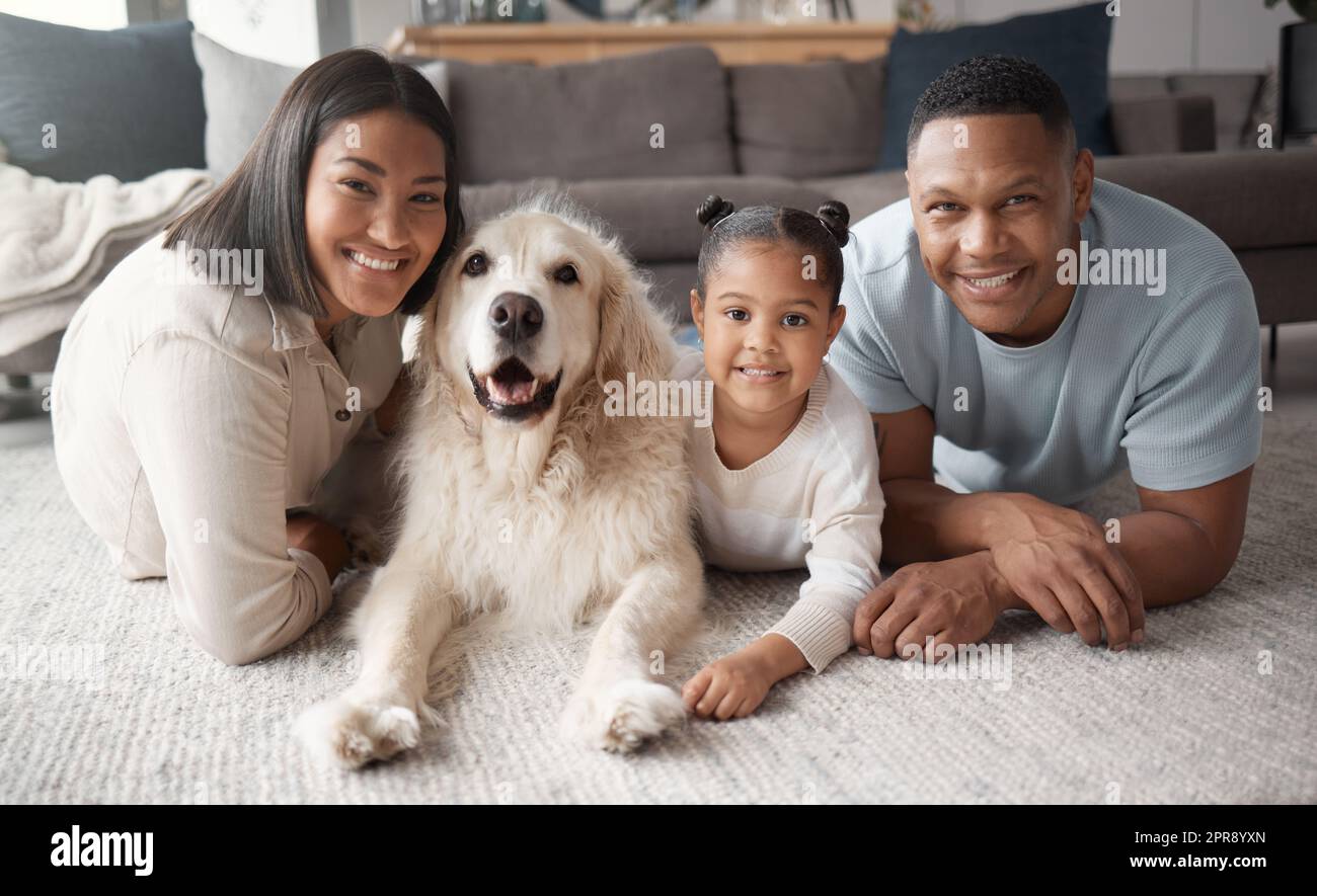 Portrait of a happy mixed race family of three relaxing on the lounge floor with their dog. Loving black family being affectionate with a foster animal. Young couple bonding with their daughter and rescued puppy at home Stock Photo