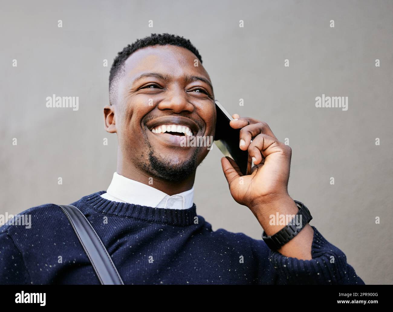 African american male on a phone call with his mobile device outside a building during the day while smiling Young black male talking on a phone while commuting to work Stock Photo
