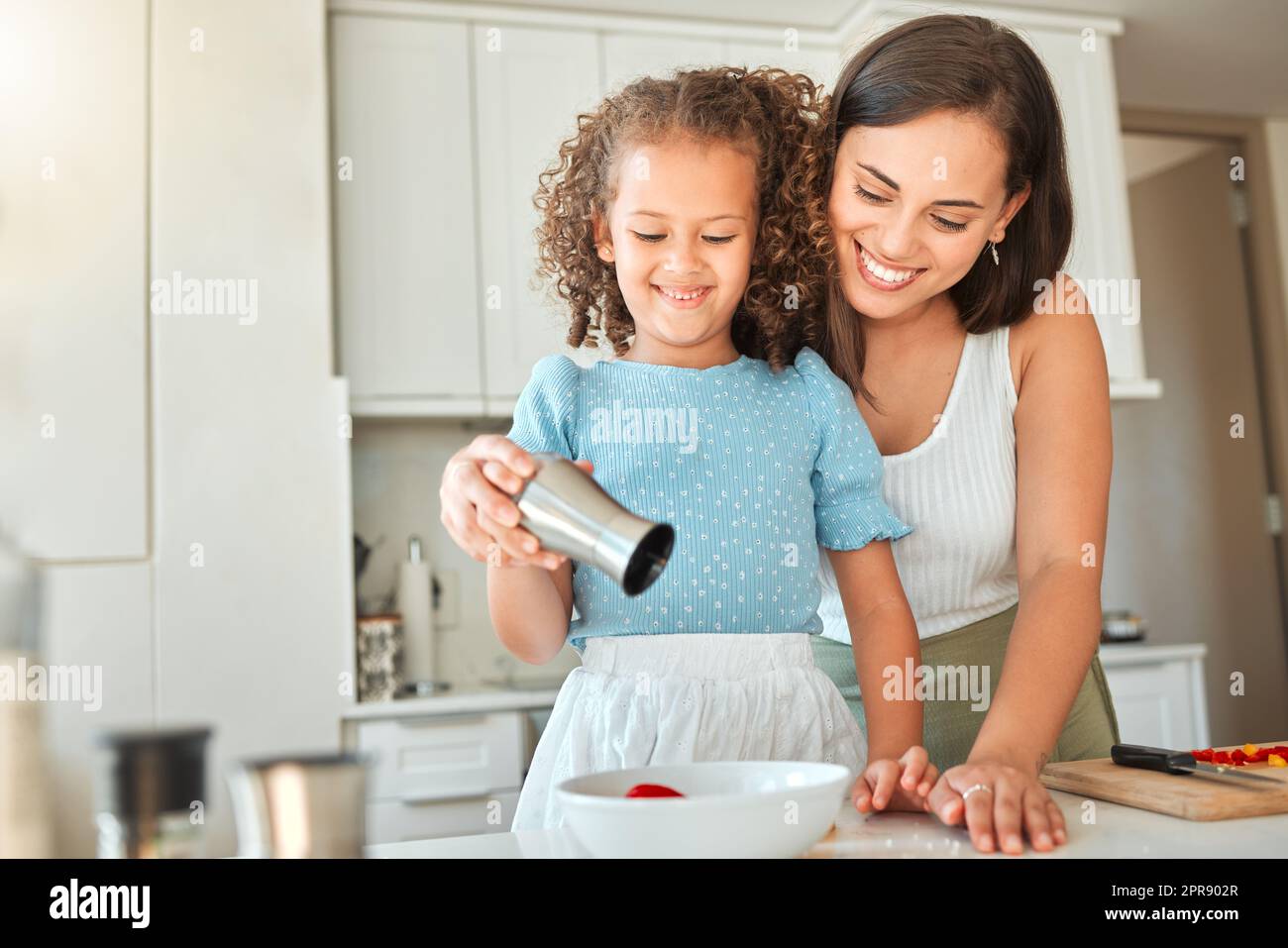 Happy mother teaching little daughter to cook in kitchen at home. Little girl adding seasoning to a bowl while making a salad with mom Stock Photo