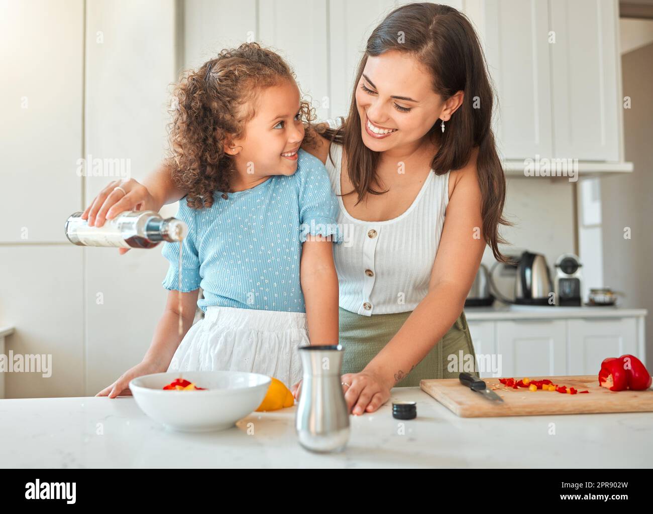 Happy mother and little daughter cooking together in the kitchen. Woman adding salad dressing while preparing a vegetarian meal with her child Stock Photo