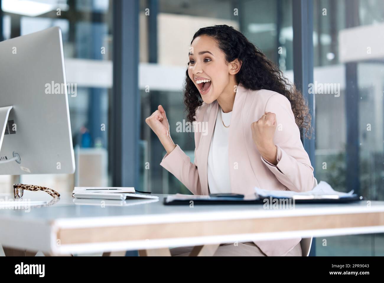 Young happy mixed race businesswoman cheering with joy using a desktop computer in an office. One cheerful hispanic businessperson smiling while looking surprised looking at a pc screen at work Stock Photo