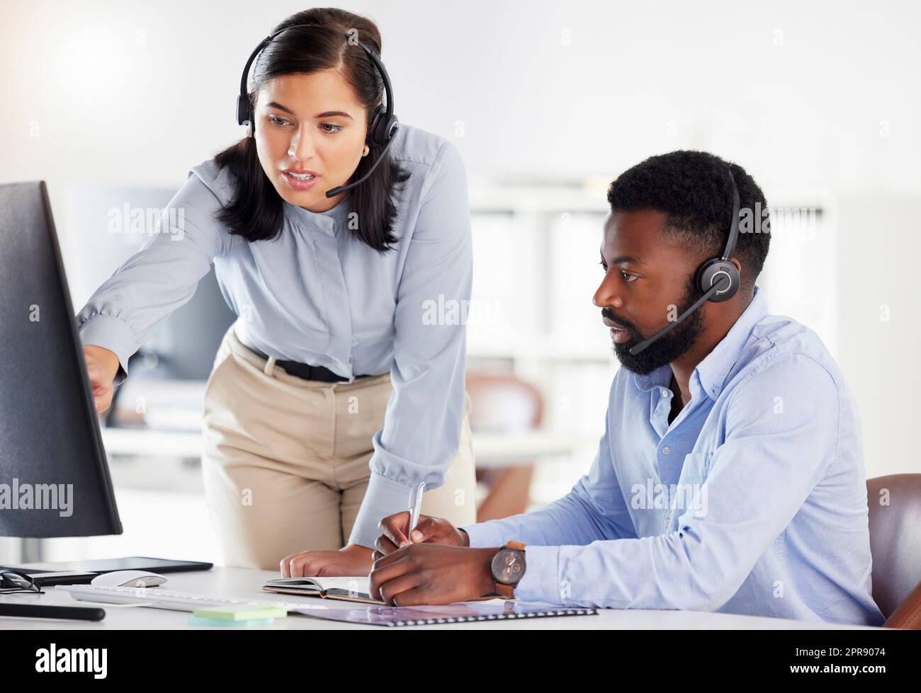 Young caucasian call centre telemarketing agent training new african american assistant on a computer in an office. Team leader troubleshooting solution with intern for customer service and sales support. Colleagues operating helpdesk together Stock Photo