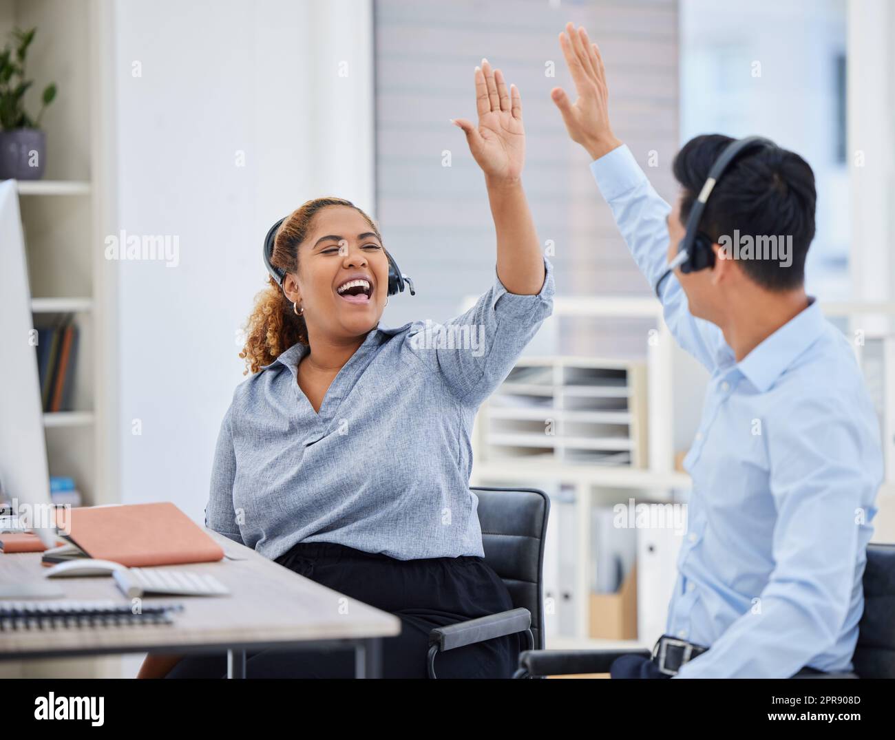 Two happy young call centre telemarketing agents giving each other a high five while cheering with joy in an office. Excited african american assistant celebrating with colleague on successful sales and reaching targets to win Stock Photo