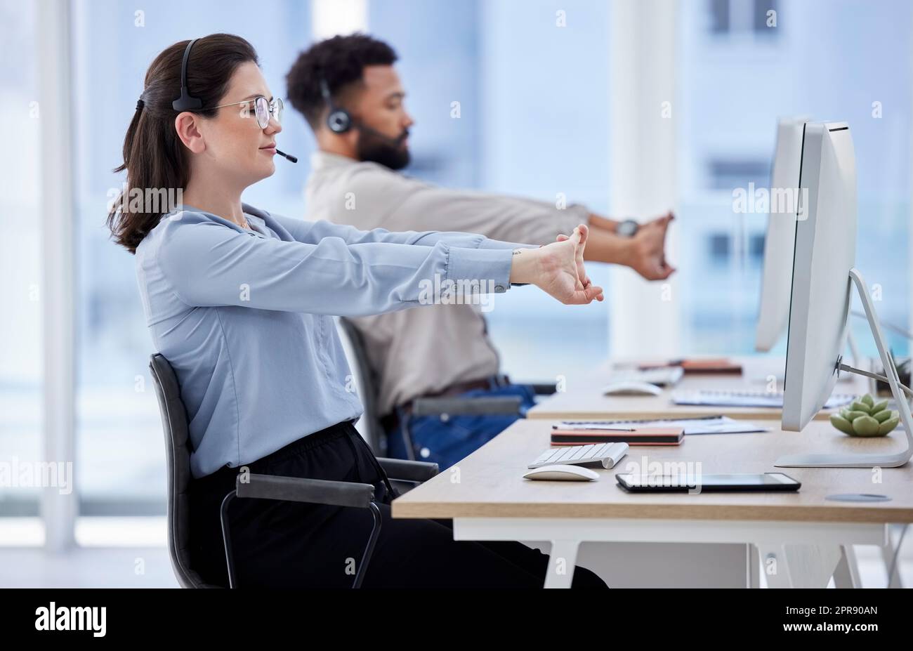 Young caucasian call centre telemarketing agent stretching her arms while working alongside a colleague in an office. Two consultants taking a break to get ready to operate helpdesk for customer service and sales support Stock Photo