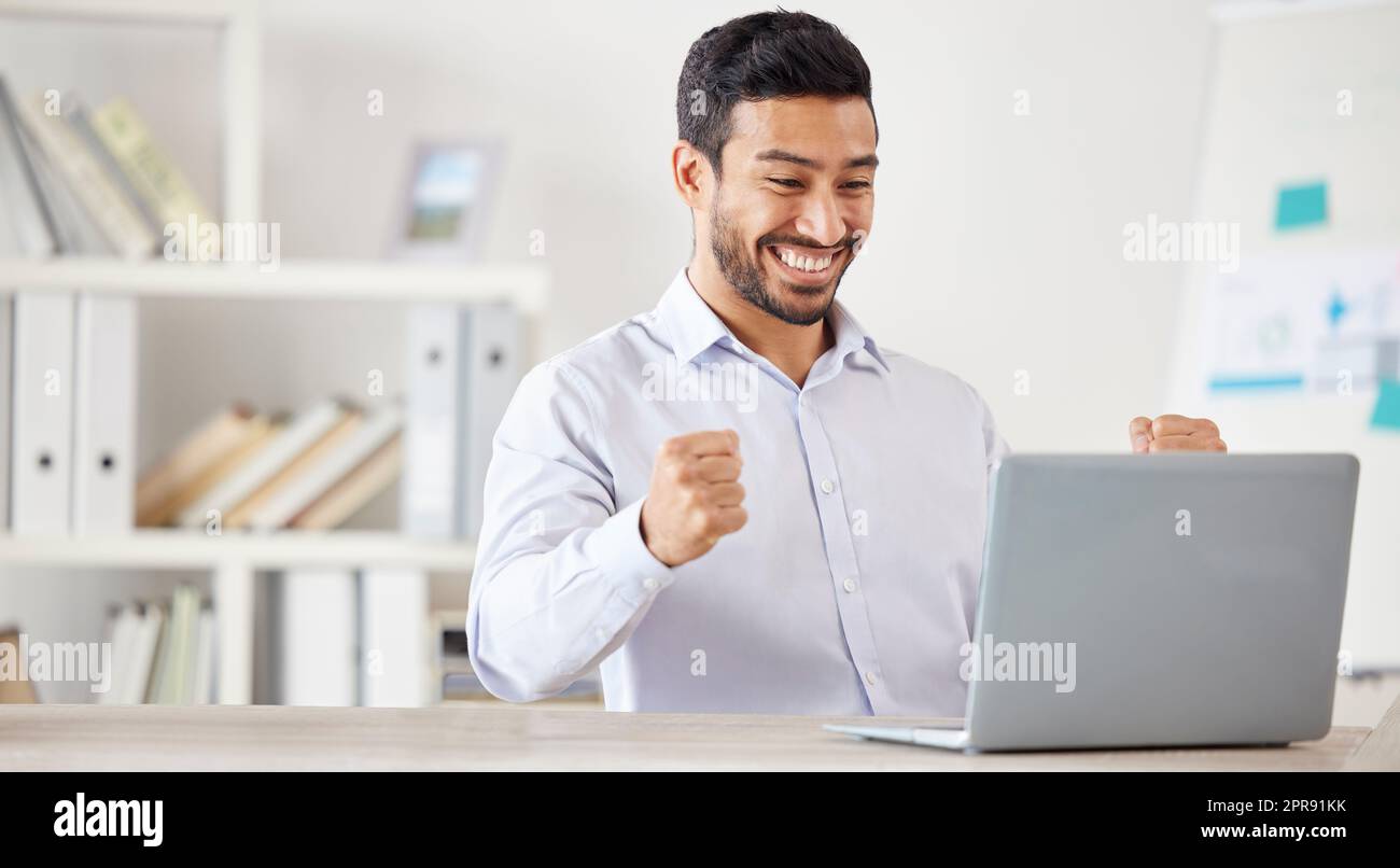 Young happy and excited mixed race businessman cheering in support while working on a laptop alone in an office at work. One hispanic male boss smiling while celebrating success and victory Stock Photo