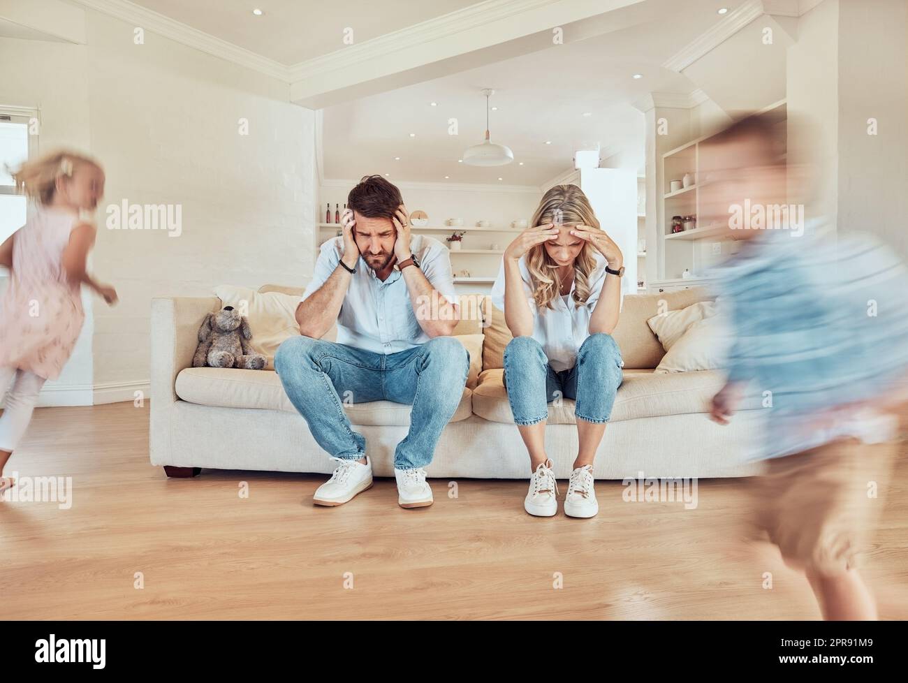 Young caucasian parents suffering from a headache while their children run in the lounge at home. Carefree playful little siblings running in the living room while their mom and dad look tired Stock Photo